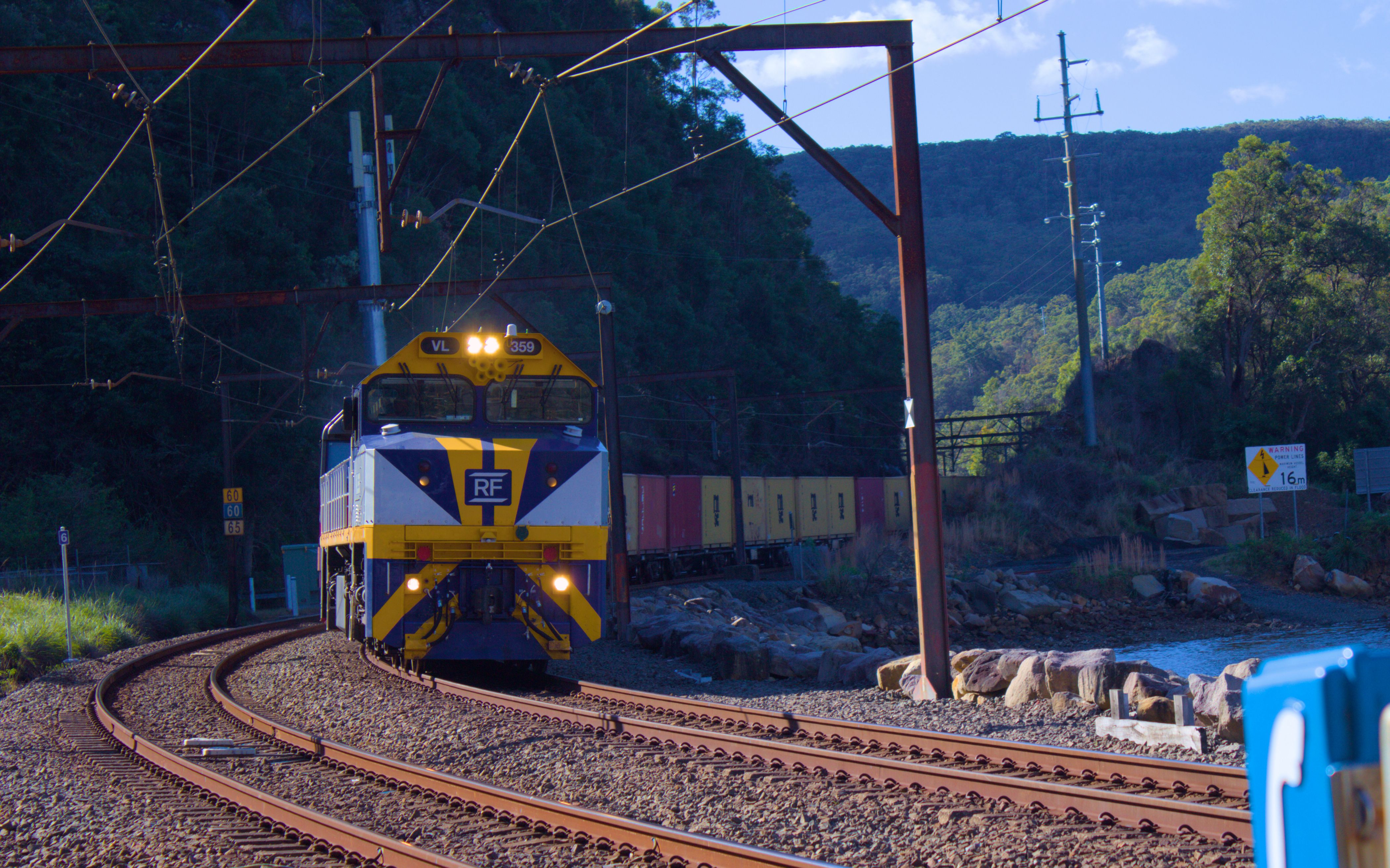 A white, dark blue and yellow freight locomotive with the lettering 'RF' on the front of it leads a long container train around a curve, with yellow and red shipping containers visible behind the train. The track is on a low causeway with a creek visible to the right of the frame. The front of the train is just passing under a rusted overhead wiring gantry. Green forested hills are visible in the background.