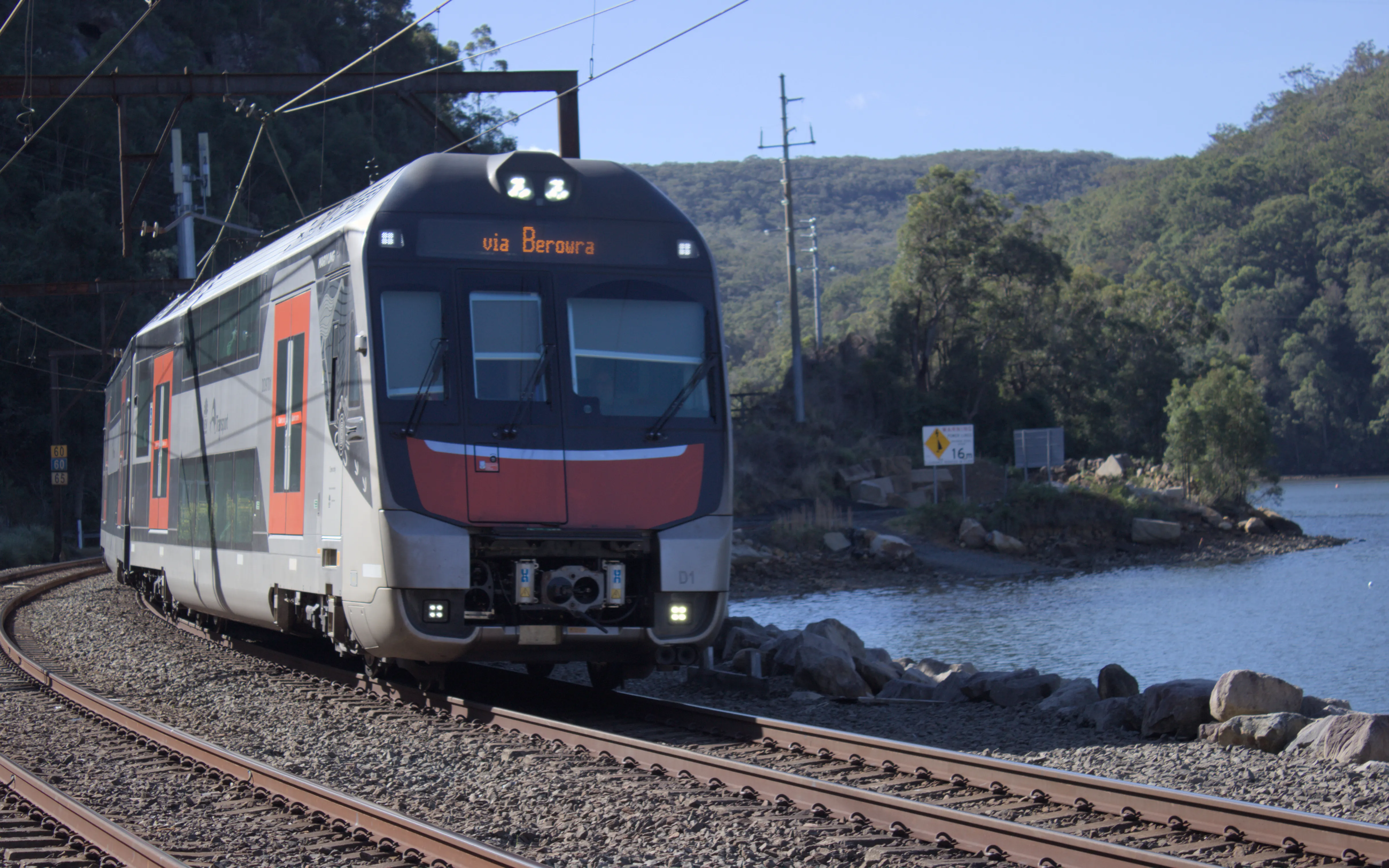 A new orange, silver and black double decker "Mariyung" intercity train  traveling along tracks on a causeway next to Mullet Creek. The tracks are  curved, with only the first and a bit of the second carriage visible as the  train is approaching the camera on the further of two tracks. Overhead wires  are visible and a rusted gantry can be seen behind the train. The background  is forested and there are hills. The sky is a dull clear colour.