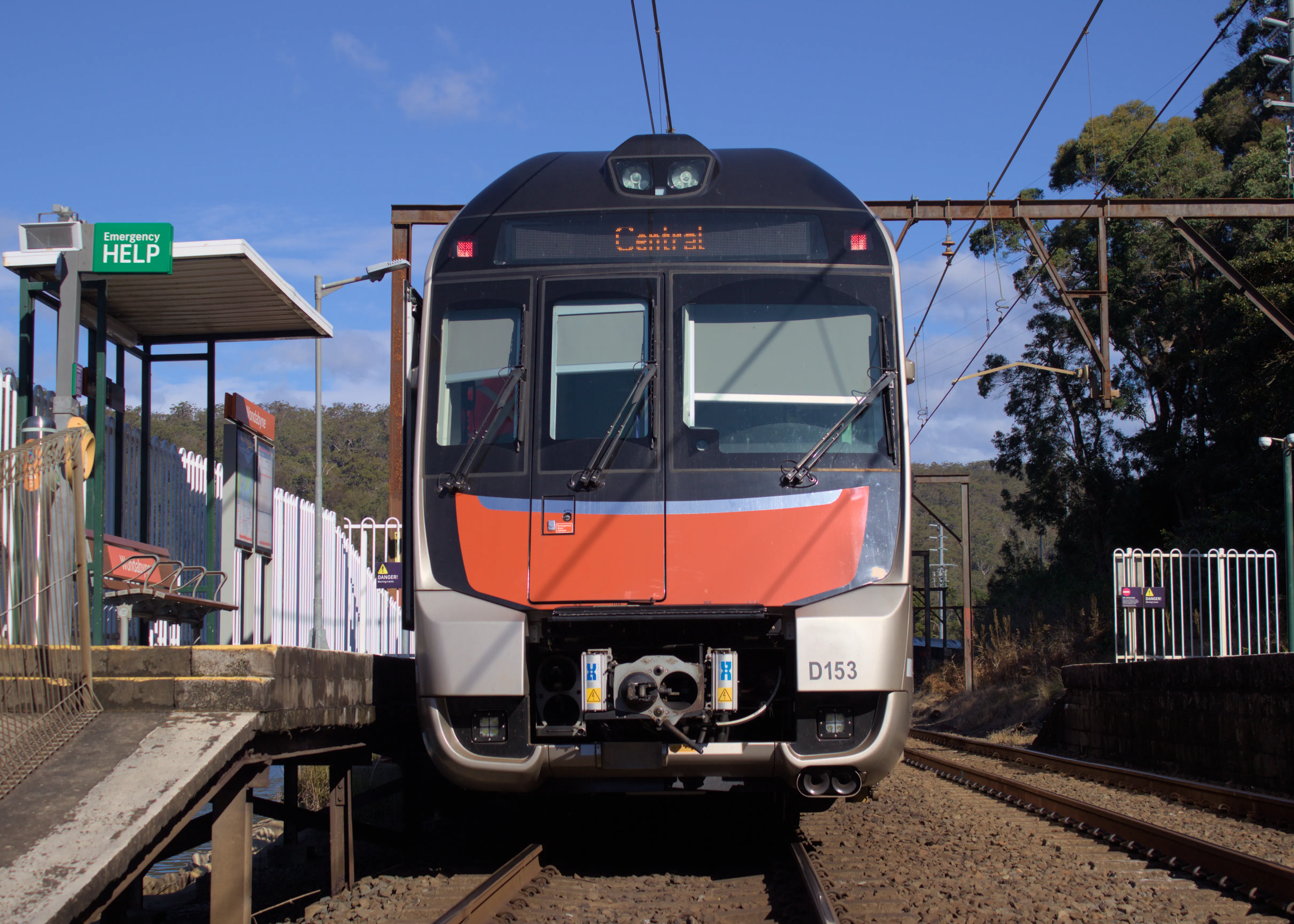 The orange, black and silver rear of a double-decker Mariyung intercity train stopped at a short concrete platform. The train's electronic destination board above its clean windshield reads "Central". This photo is taken at a low angle on the tracks, looking up at the train. The train is on the left track, while the right track and other platform is unoccupied. The platform has a small shelter, a wooden bench and a fence along a ramp. The sky is mostly clear and the background appears densly forrested.
