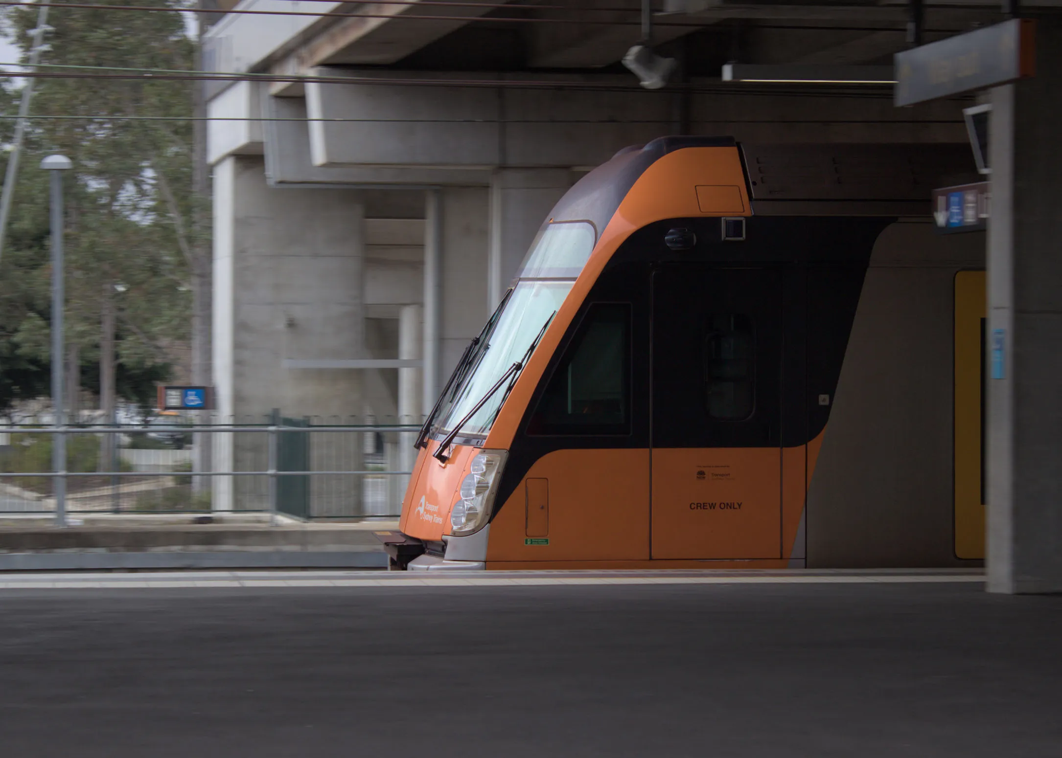 The side of the cab of a modern orange and black train with a silver body moving along a platform at speed as indicated by the blurry background.