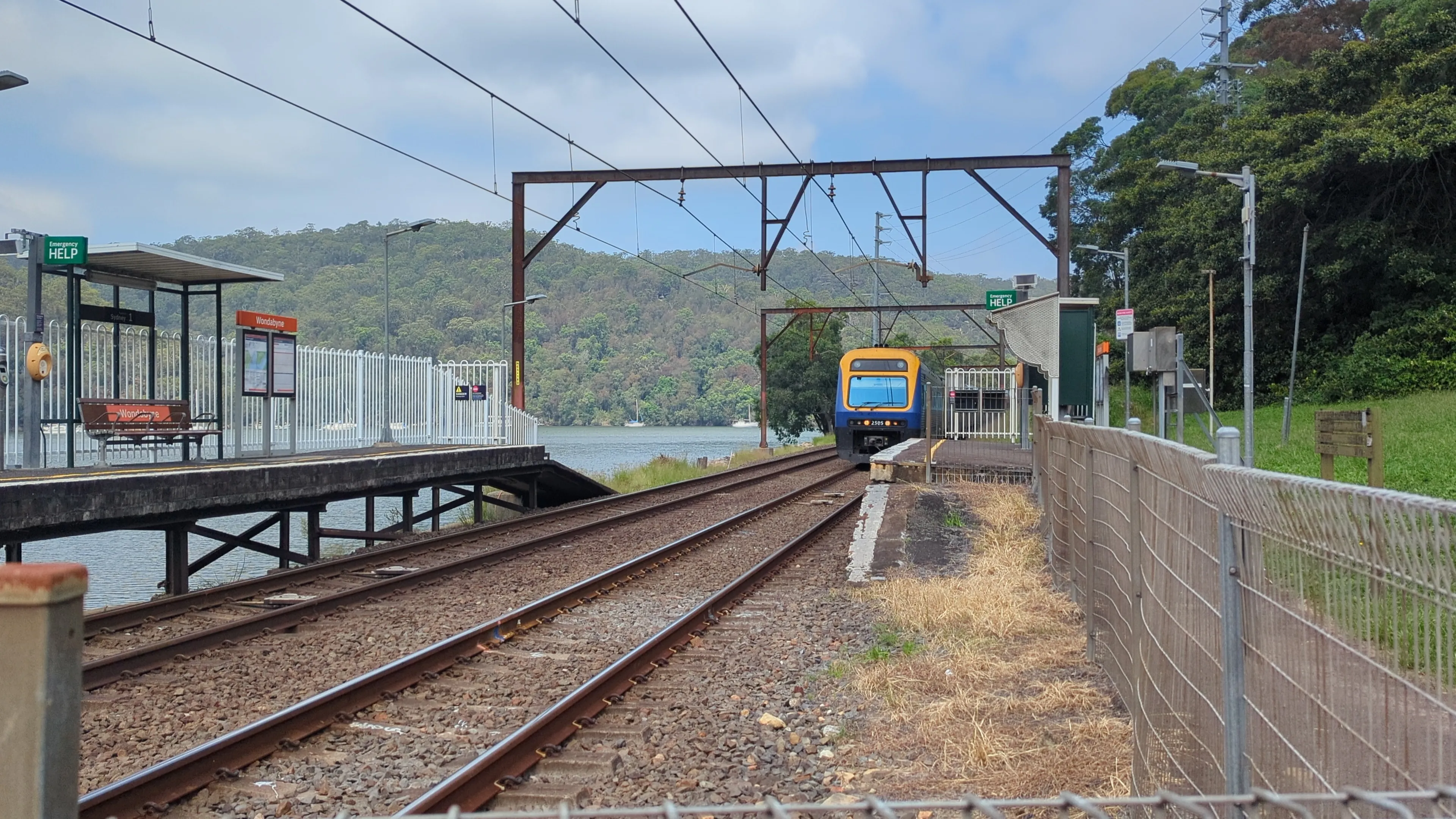 The front of a yellow and blue Xplorer train can be seen entering the small station of Wondabyne, passing under a wire gantry. It is a bright sunny day  and green hills are visible in the background along with Mullet Creek.