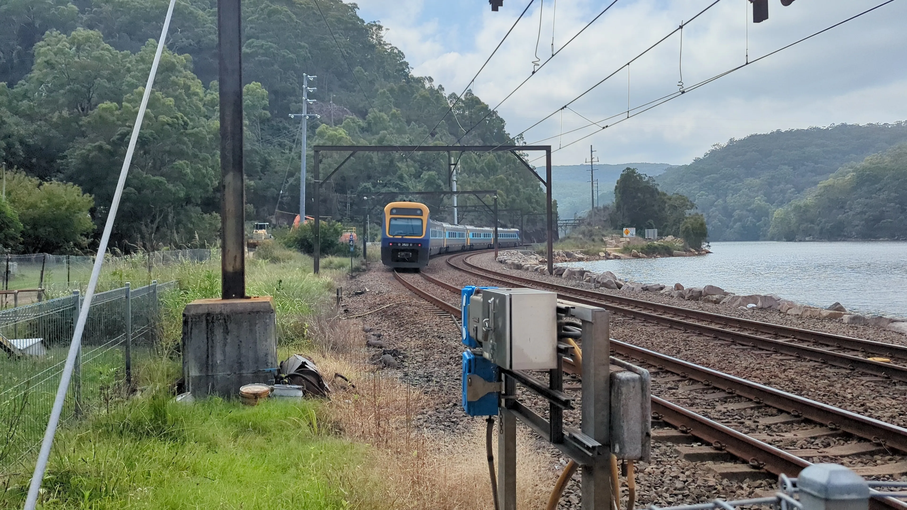 A 5-carriage long blue, yellow and silver Xplorer train heading around a curve in the tracks past Wondabyne, with Mullet Creek visible to the left of the causeway. The sky is partly cloudy and there is abundant greenery in the background.