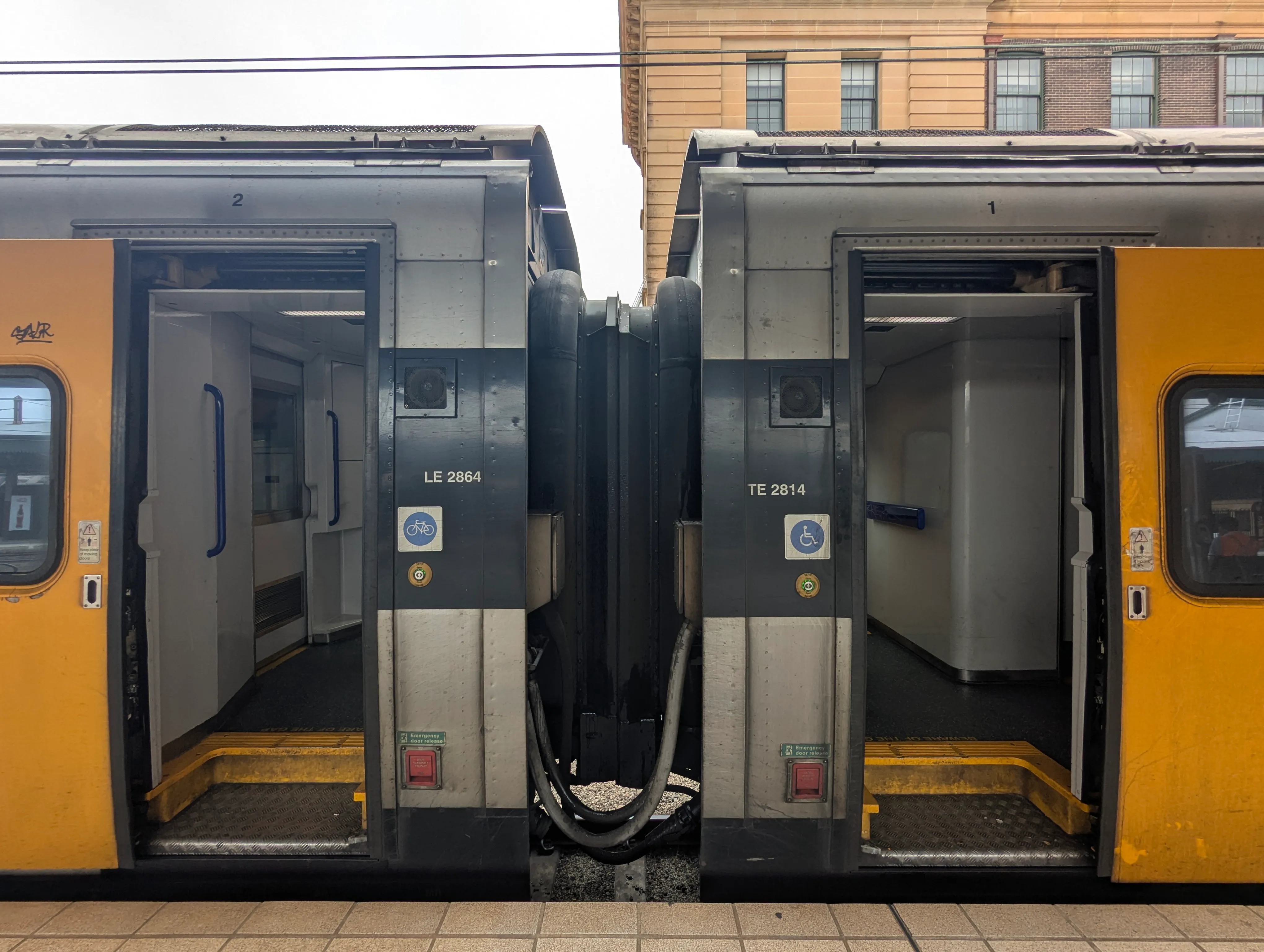 Two connected train carriages side-by-side at a station platform. Both doors  are open, showing the interior of the train. There are accessibility symbols  next to the doors, with the one on the left indicating bicycle access and the  other wheelchair access. The door is yellow, whilst the rest of the train is  silver-grey. The top of a building is visible in the background, made of brick. The image is taken during daytime, and the sky is overcast.