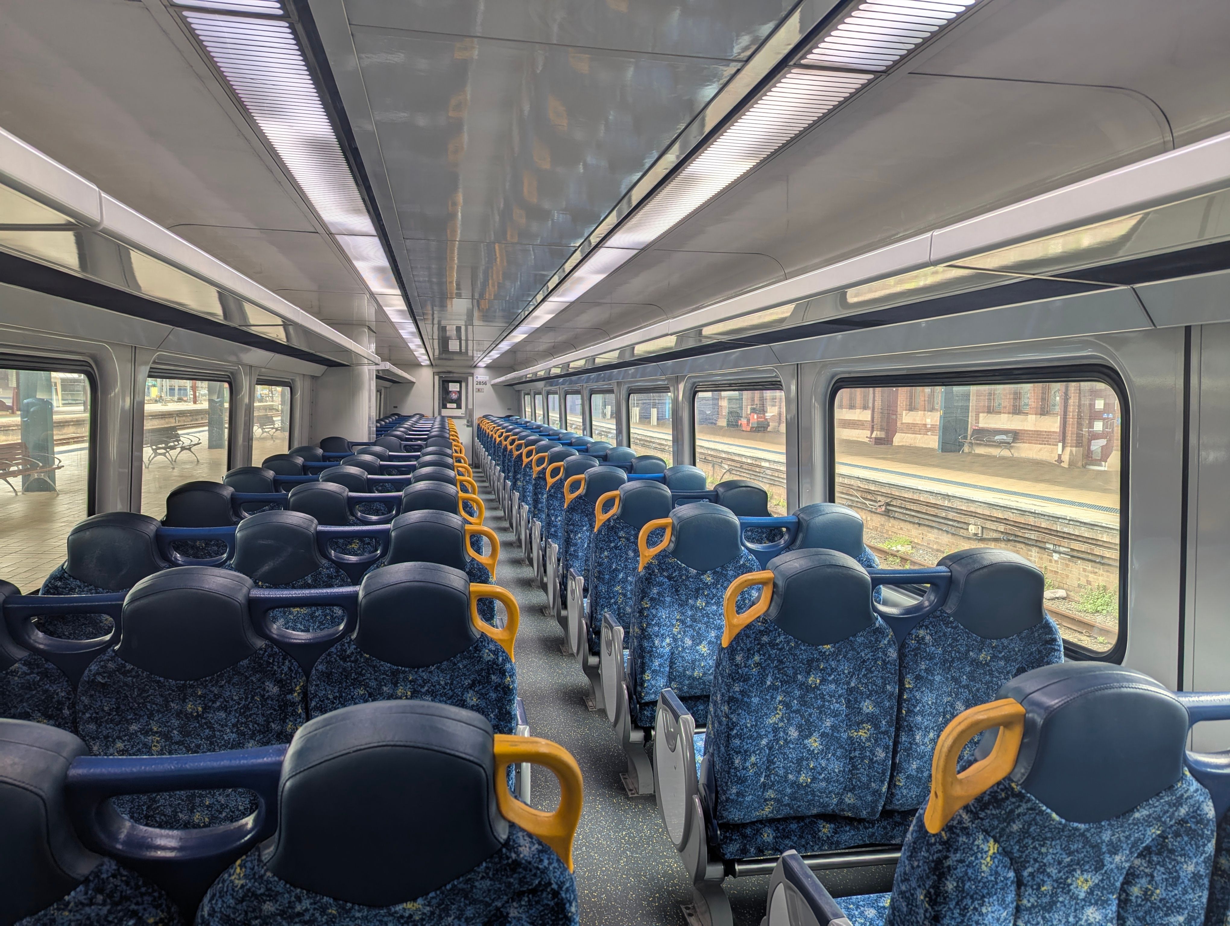 The interior of the train. It is clean and brightly lit, with white walls and ceiling and reversible blue-patterned fabric chairs in a 3-2 configuration. The chairs have a headrest and yellow handles on aisle seats. Large clear windows line both sides, providing views of the station platform outside. The station appears empty, and natural daylight fills the carriage.