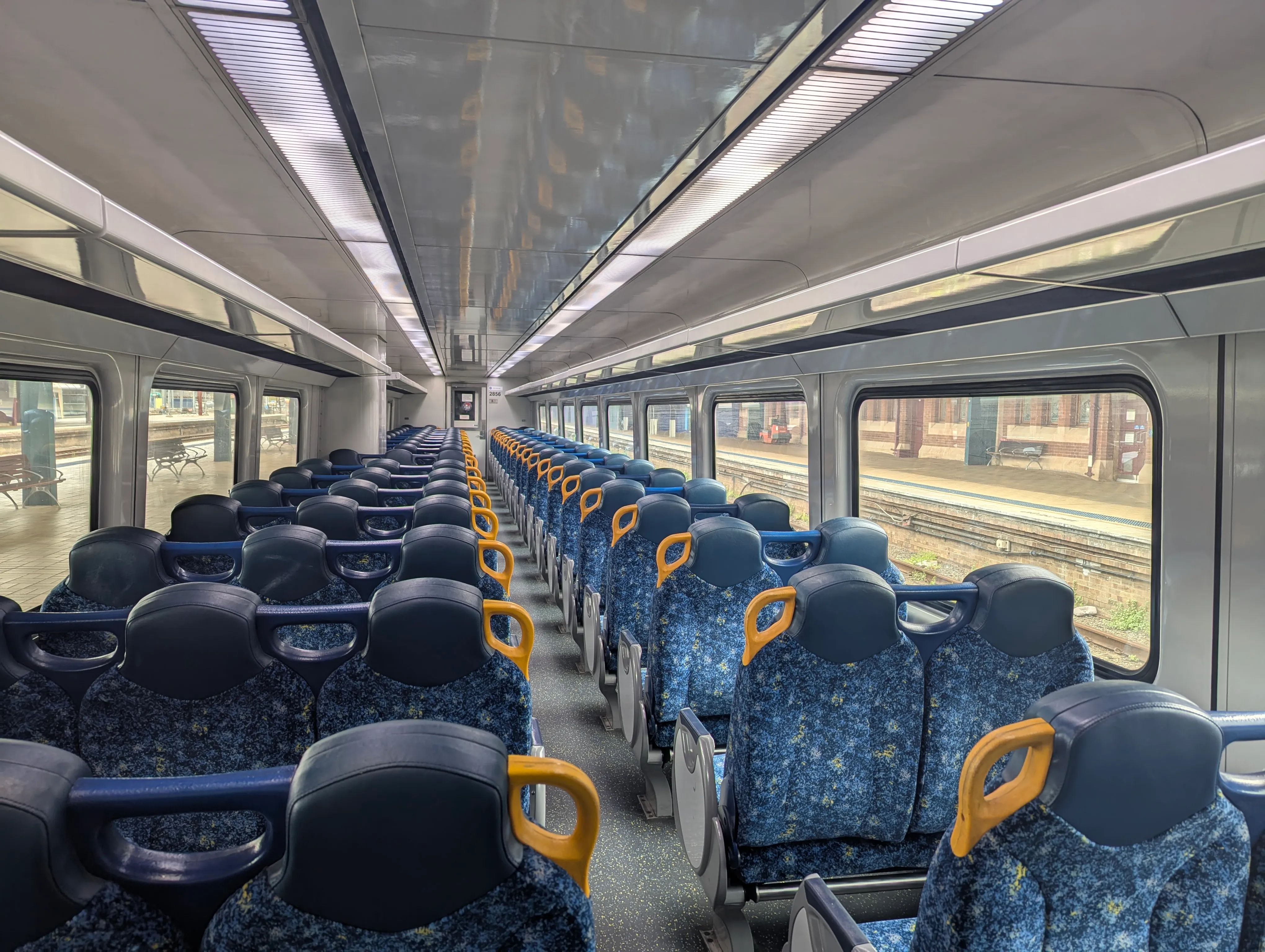 The interior of the train. It is clean and brightly lit, with white walls and ceiling and reversible blue-patterned fabric chairs in a 3-2 configuration. The chairs have a headrest and yellow handles on aisle seats. Large clear  windows line both sides, providing views of the station platform outside.  The station appears empty, and natural daylight fills the carriage.