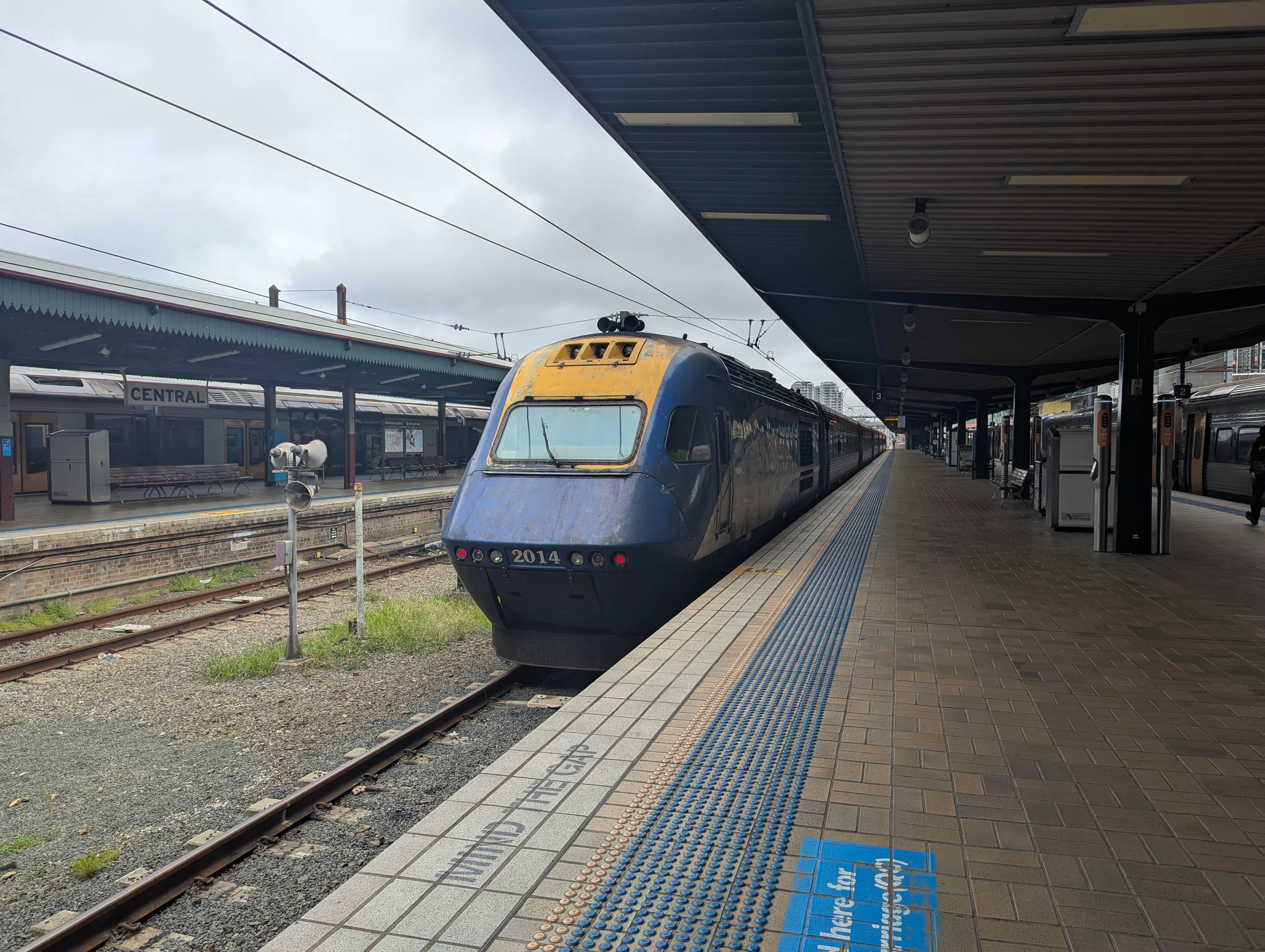 This image shows a blue and yellow power car at a station platform. The front of the power car is prominent, with a streamlined design, albeit a bit dirty. The nose of the train is curved, with the number "2014" painted on the front in white numbering. The train's body is primarily blue, with a lighter blue wave pattern running from the middle to the rear. The platform is visible in the foreground, with square tiles arranged in a grid pattern. In the background, a dark-colored train compartment can be seen. The sky above is slightly overcast.