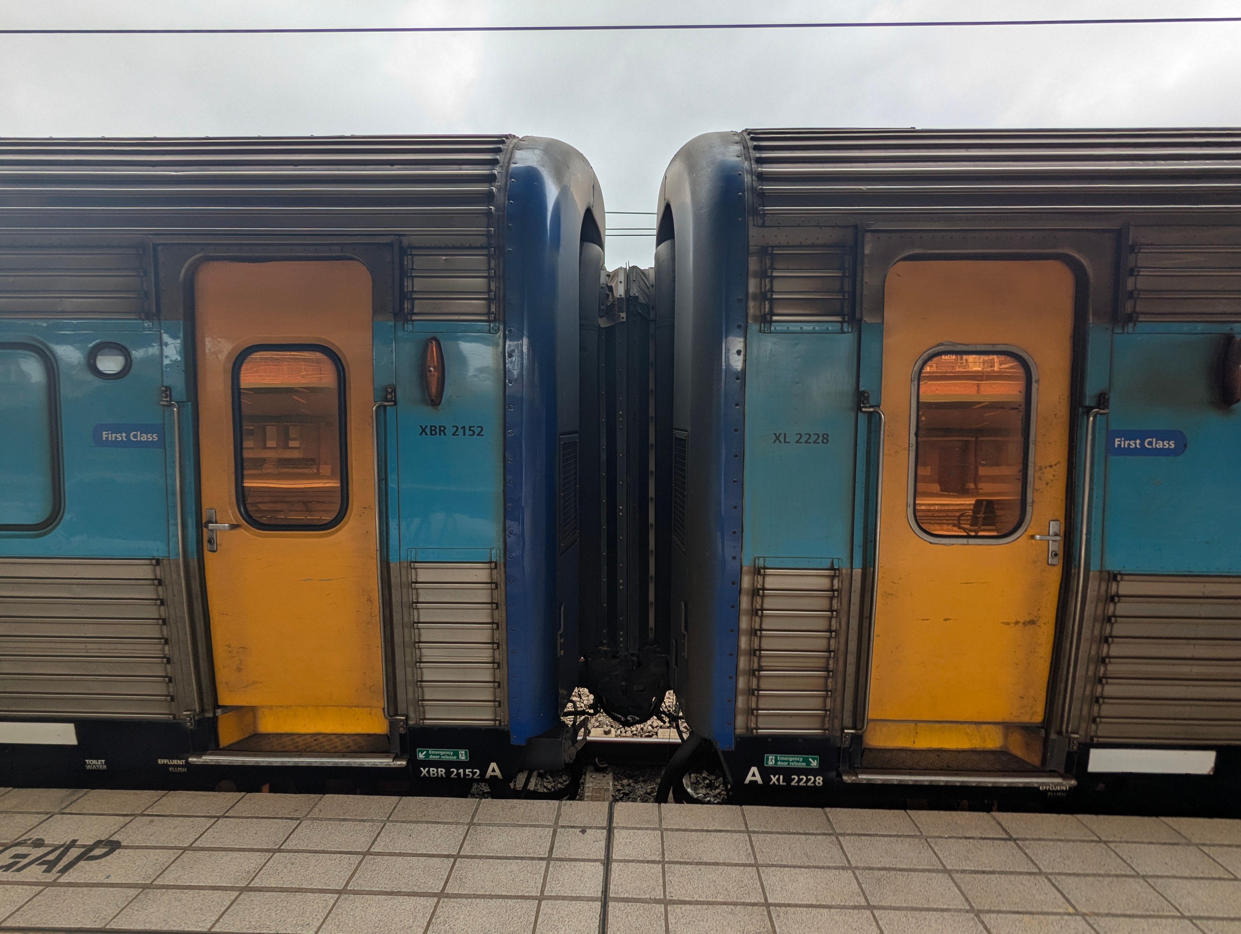 Two connected train carriages side-by-side at a station platform. Both doors which are yellow and have a copper-tinted window are closed. The carriages are made of corrugated stainless steel with a light-blue line around window-height as part of the livery. The ends of each carriage is painted in a dark blue colour, and the connector between the carriages is black. It is daytime and the sky is overcast.