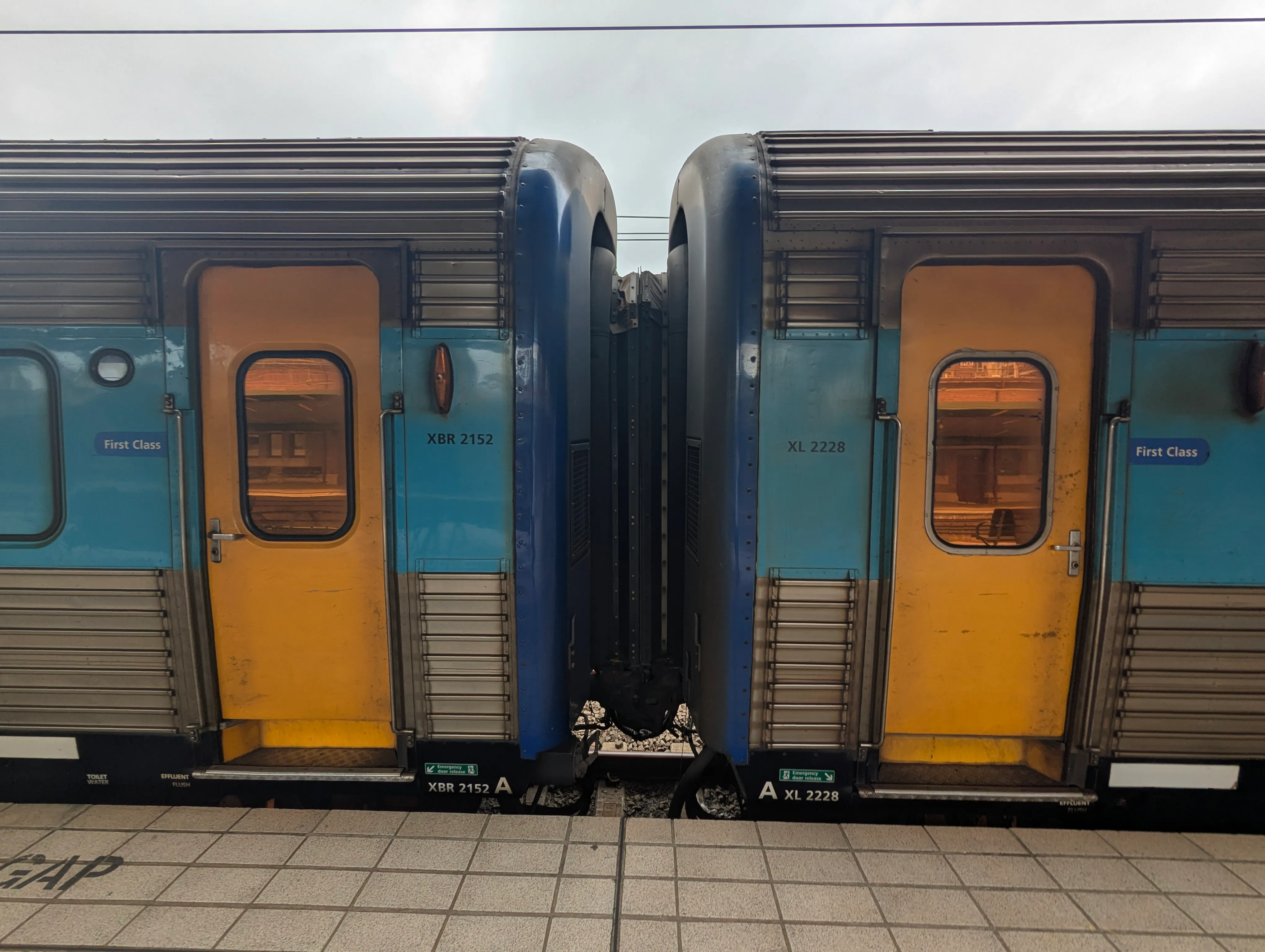 Two connected train carriages side-by-side at a station platform. Both doors which are yellow and have a copper-tinted window are closed. The carriages are made of corrugated stainless steel with a light-blue line around window-height as part of the livery. The ends of each carriage is painted in a dark blue colour, and the connector between the carriages is black. It is daytime and the sky is overcast.