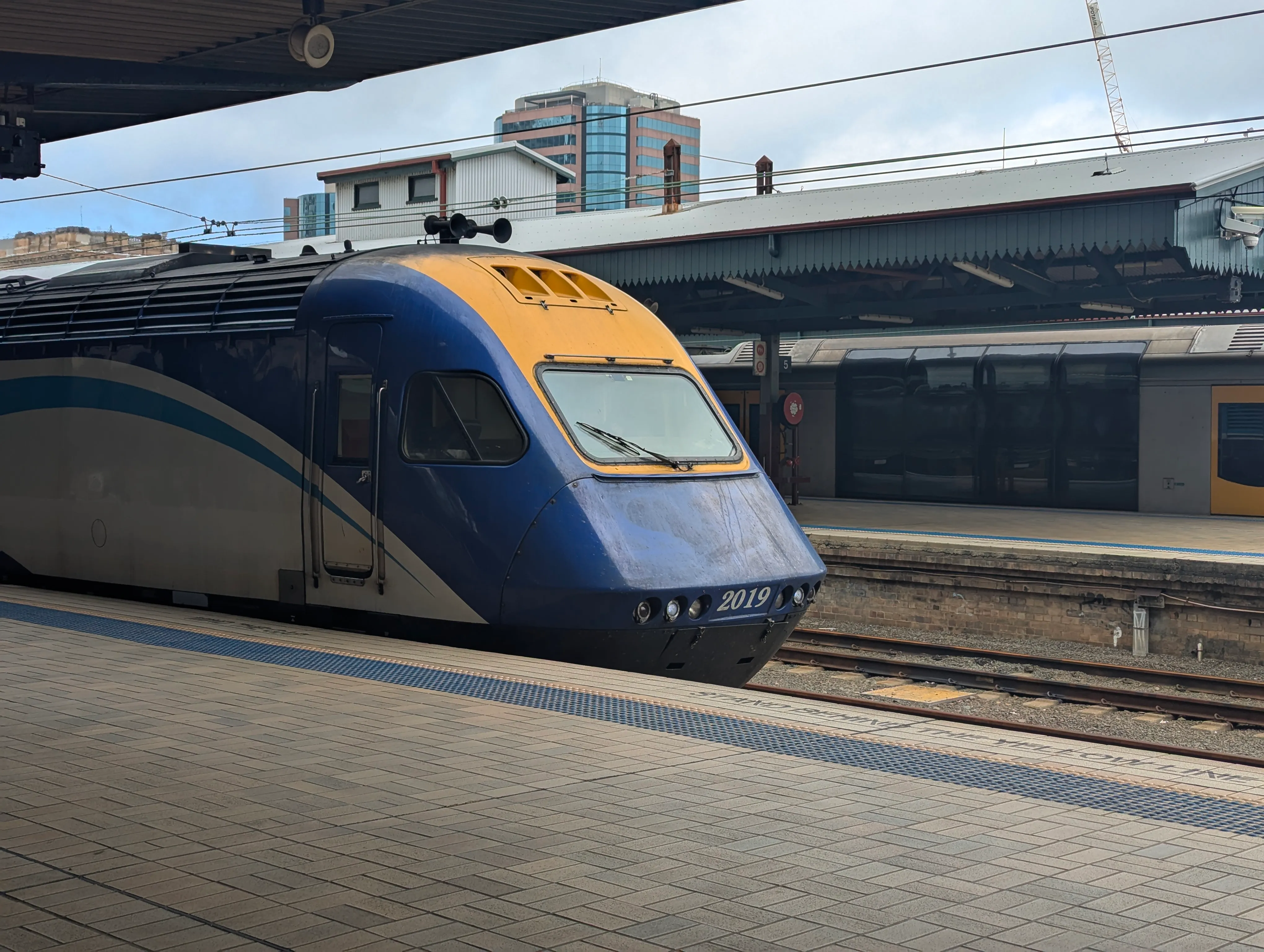 This image shows a close-up of a blue and yellow long train at a station. The front of the train is prominent, with a streamlined design. The nose of the  train is curved, with the number "2019" displayed on the front in white. The  train's body is primarily blue, with a lighter blue wave pattern running from  the middle to the rear. The platform is visible in the foreground, with square tiles arranged in a grid pattern. In the background, a dark-colored train  carriage of a Tangara can be seen. The sky above is slightly overcast, with some  buildings visible in the background. The overall atmosphere appears to be a  busy, modern train station.