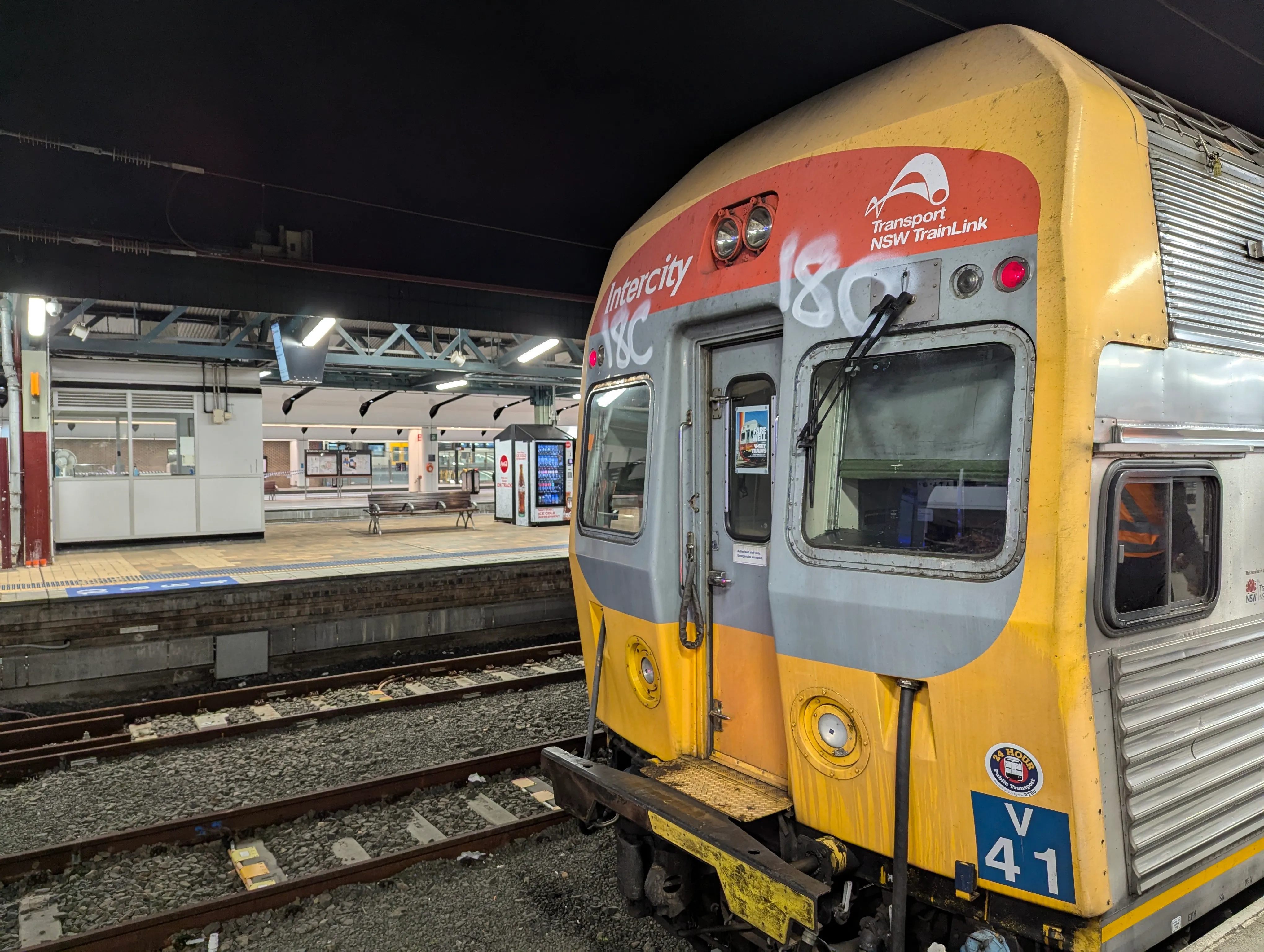 A yellow, red and grey V set on taking up the right side of the frame at the intercity platforms of Central station. The front and some of the side of the train is visible. The side of the train is made of corrugated stainless steel and is largely unpainted. On the front, the words "Intercity" and "Transport NSW TrainLink" are marked in white paint. The photo is taken at night, with an almost completely dark sky whilst the platforms are brightly lit by the station lights. 