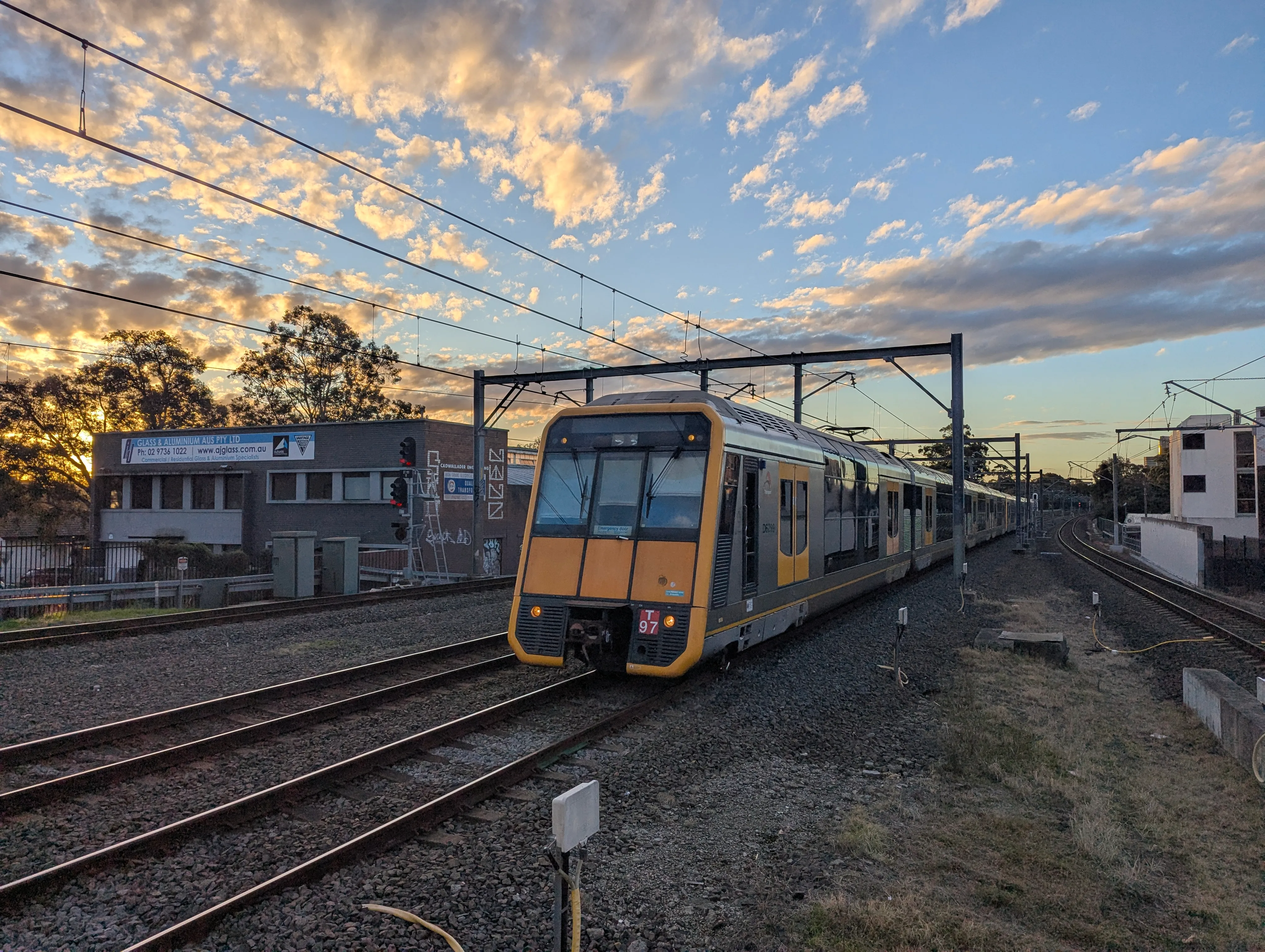 A steel double-decker 8-car suburban "Tangara" train with a yellow and black  front, and yellow doors passes under an overhead wire gantry on the centre  right track out of four tracks. The red target plate on the front of the train indicates T97 while the destination board is empty, presumably broken. Some  short buildings can be seen to the left and right of the rail corridor with  some light graffiti. The sky is a light blue colour with some clouds and the  lighting is that of a sunset from the left.