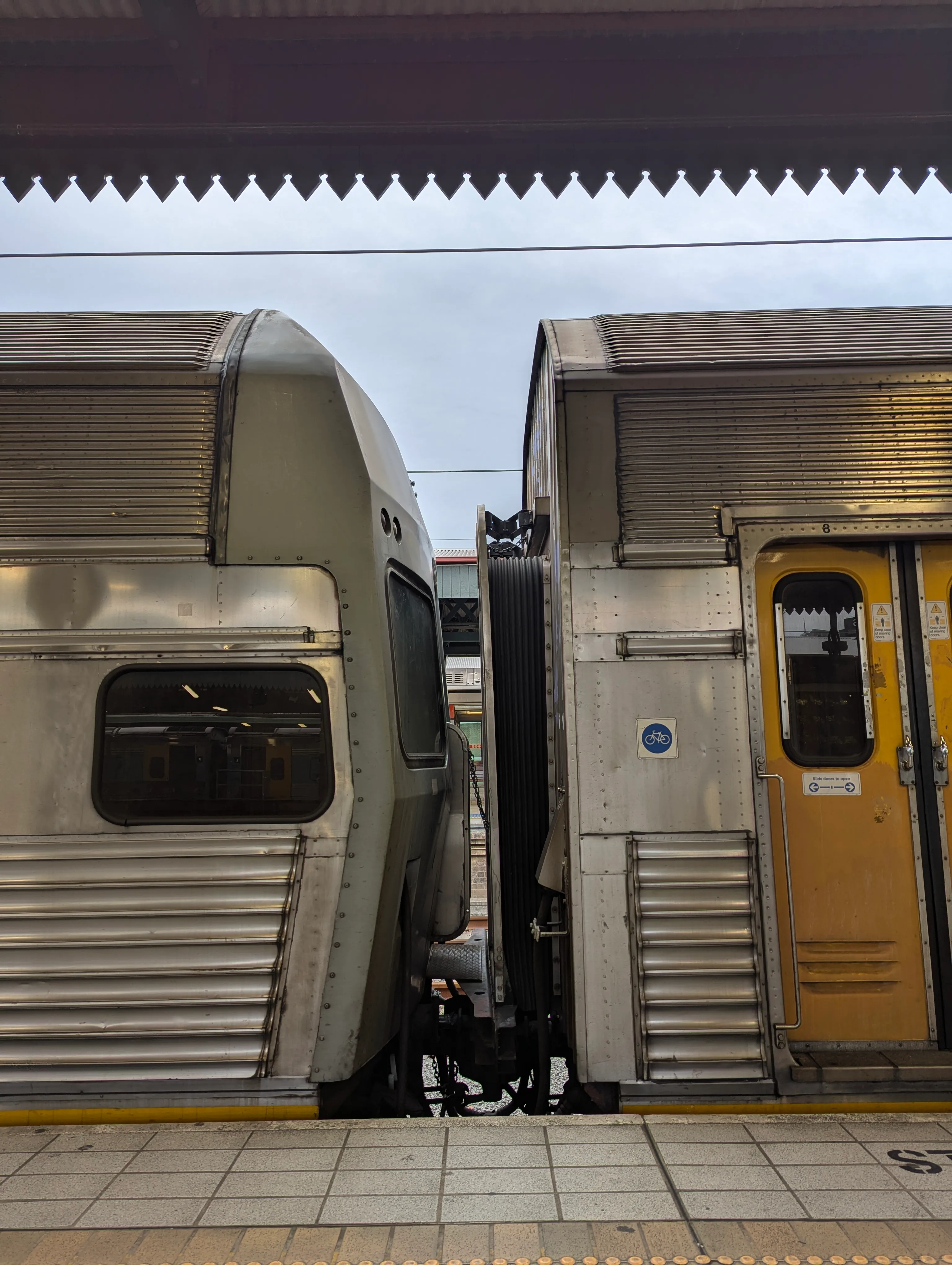 Two fluted silver-bodied train carriages attached to each other, with the coupling and partially open gangway visible at the centre of the frame (portrait orientation). The door on the right carriage is visible and is yellow. The sky is overcast and the roof of the platform is visible to the top of the frame. 