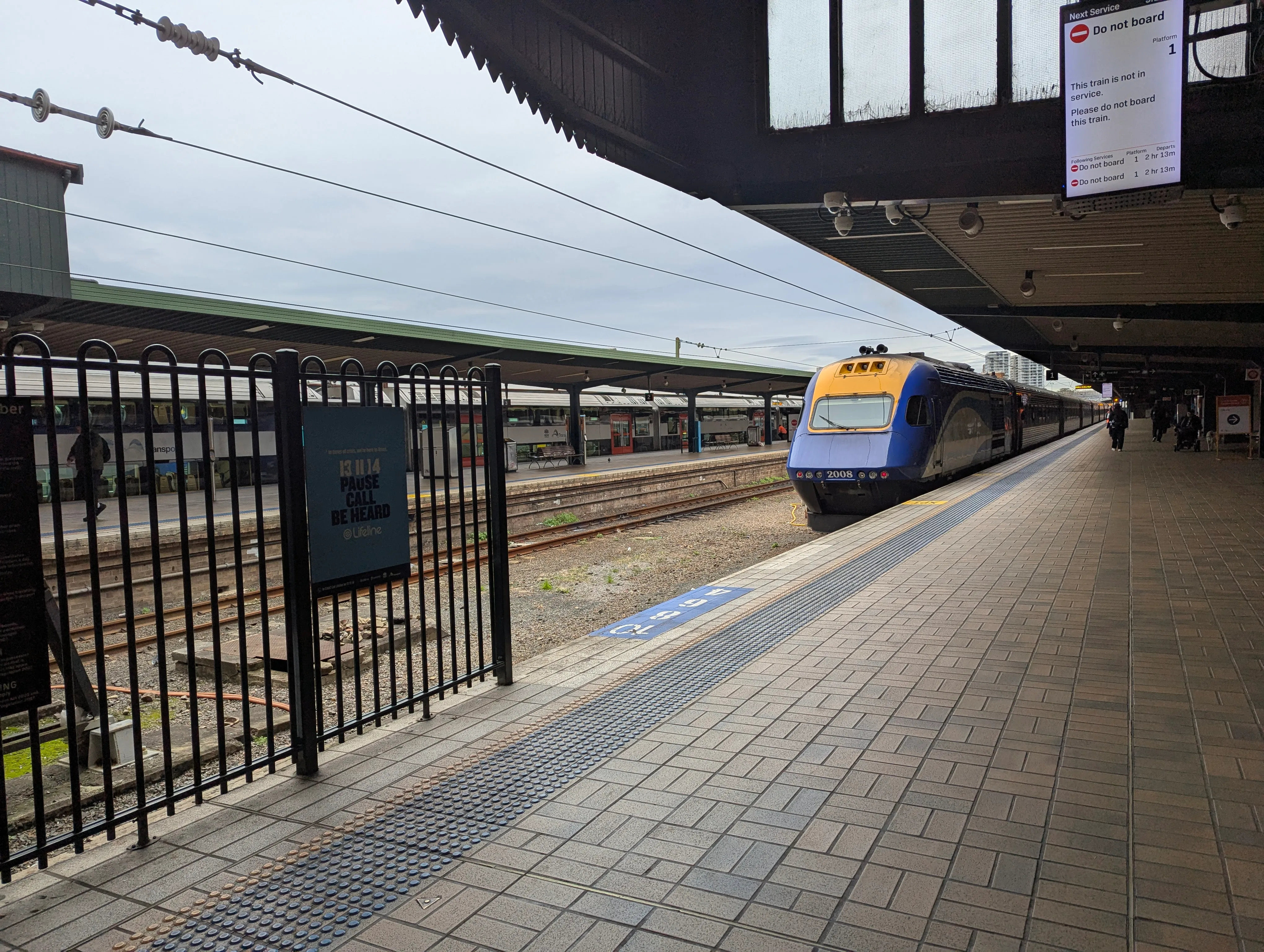 A blue, yellow and white liveried XPT power car "2008" sitting at the platform with six passenger cars visible behind it. A display board mounted to the roof of the platform informs passengers "Do not board" as "This train is not in service." The sky is overcast and the lighting is even.