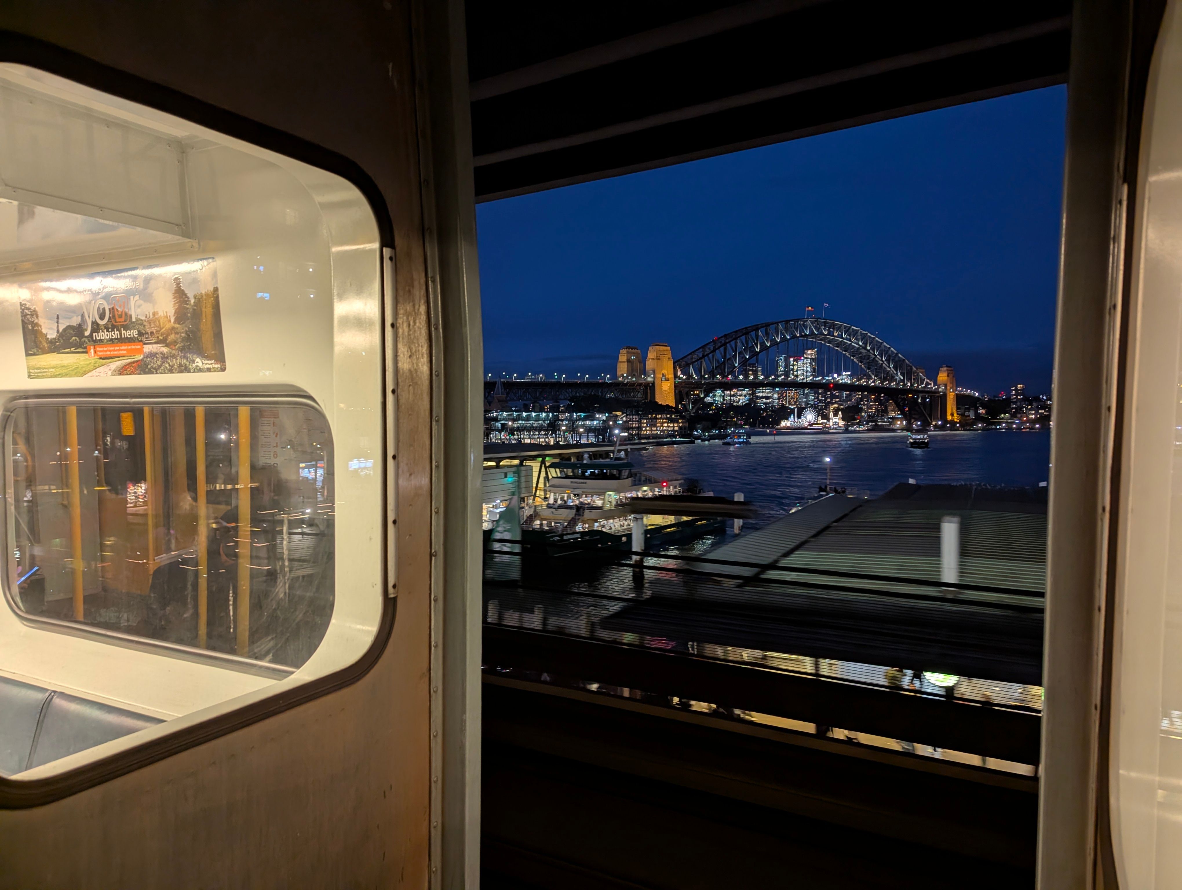 A shot taken between two carriages of a silver K set at night with the iconic Harbour Bridge lit up visible toward the centre of the frame on a dark blue  sky. Circular Quay is visible to the botttom of the shot.