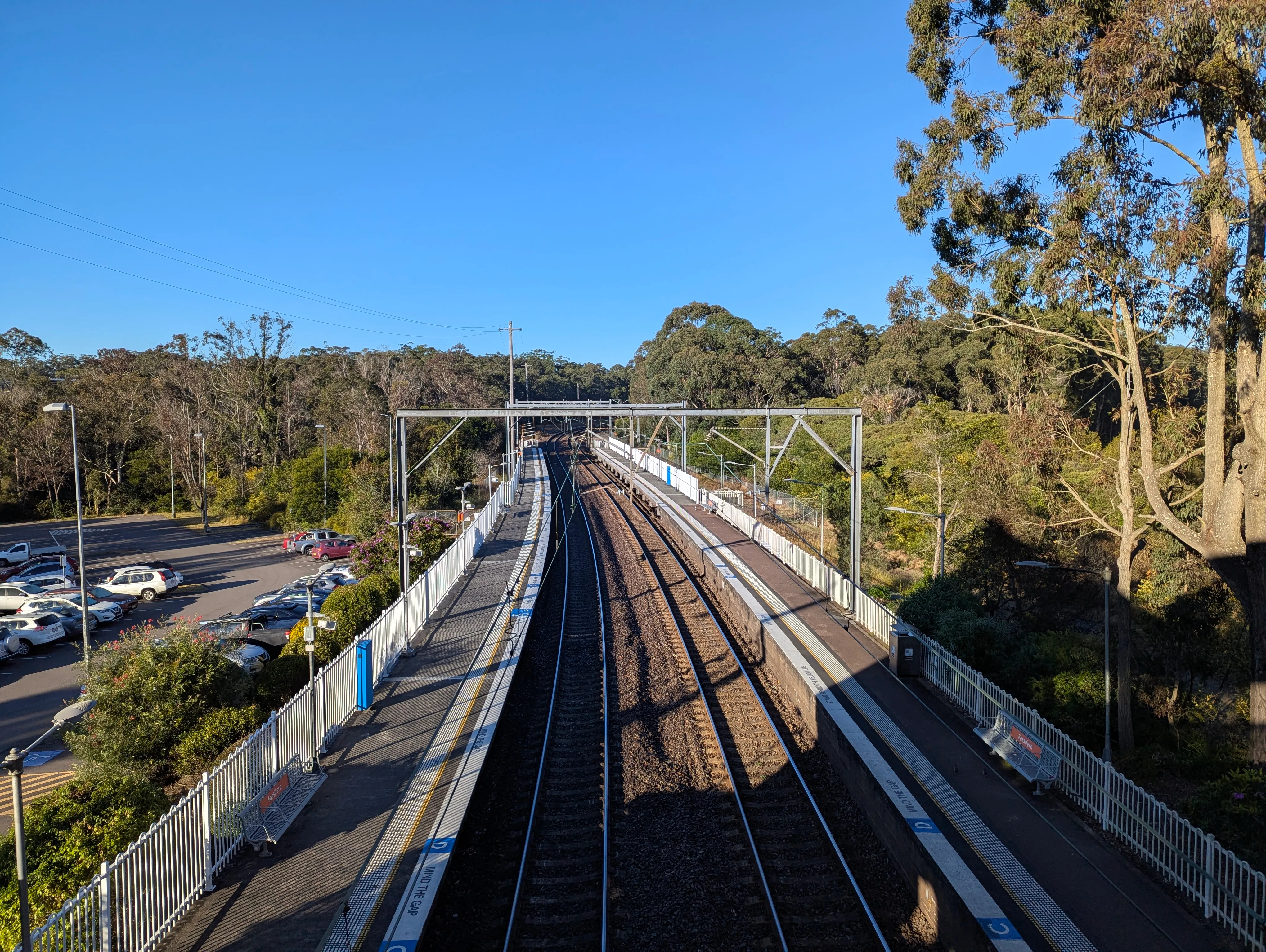 Looking over a station from a footbridge, a pair of tracks are visible that  extend into a curve in the distance with a pair of platform on either side. Steel wire gantries are visible at regular intervals on along the platform. There is a small commuter carpark visible on the left outside the station, and the whole place is surrounded by trees and bushland. The sky is a clear blue colour. 