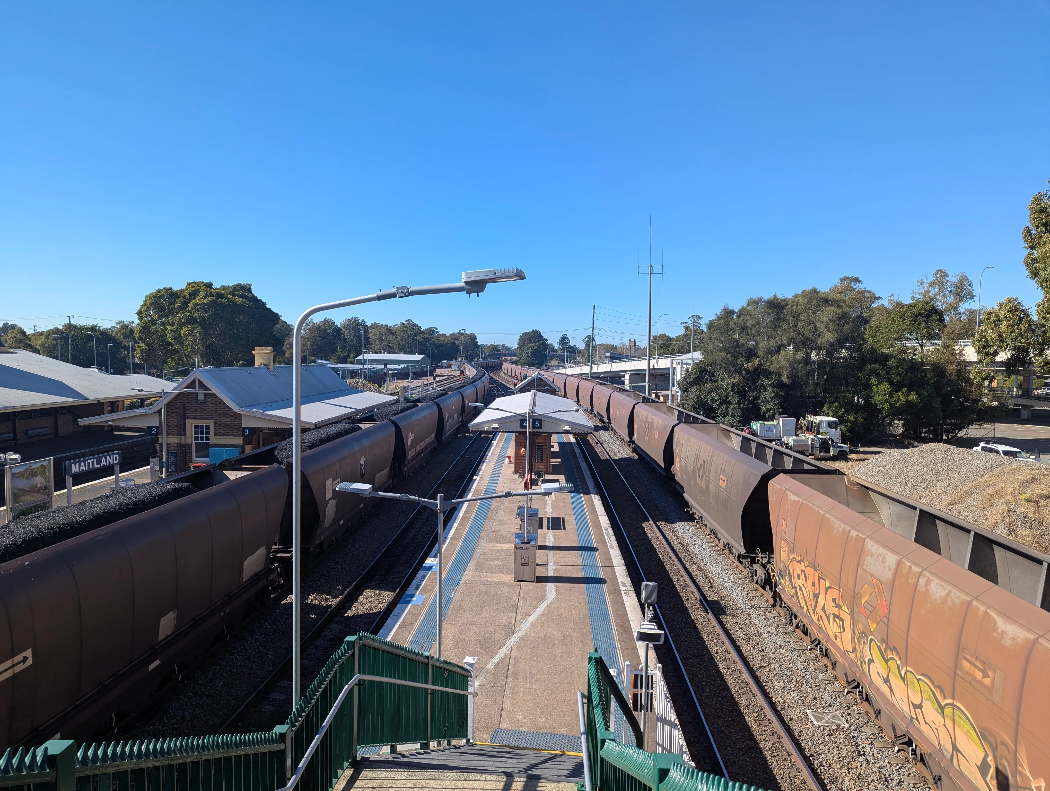 Two very long trains made of coal hoppers passing each other, with a platform visible in the middle of the frame. The train on the left is moving away from the camera with hoppers full of coal while the train on the right is moving toward the camera with empty hoppers. The sky is bright and clear. 