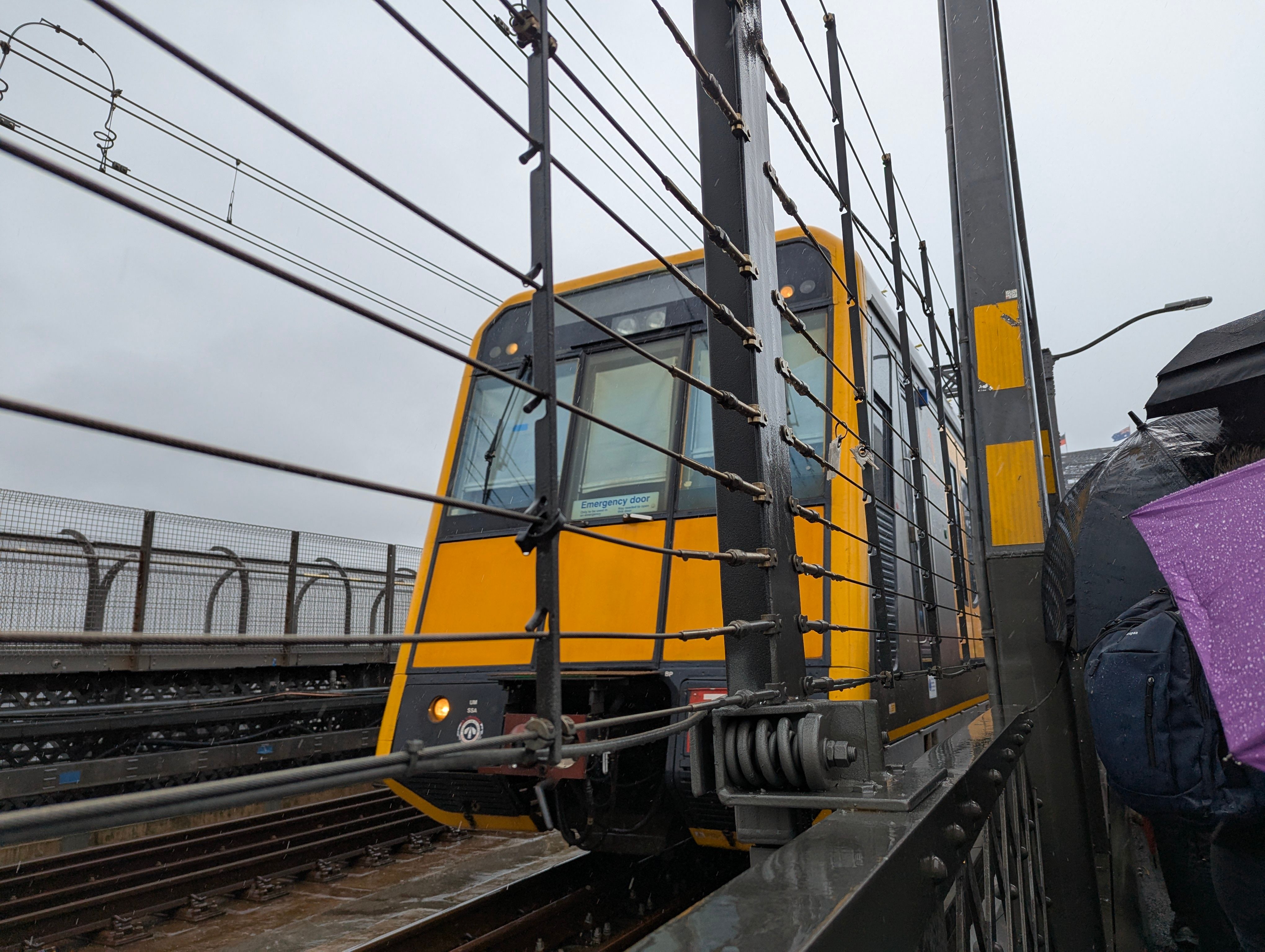 A low-angle close up shot of the distintive slanted yellow front of a suburban "Tangara" train with a wire fence visible between the camera and the train. The sky is a dull white colour and the train and surroundings are wet.