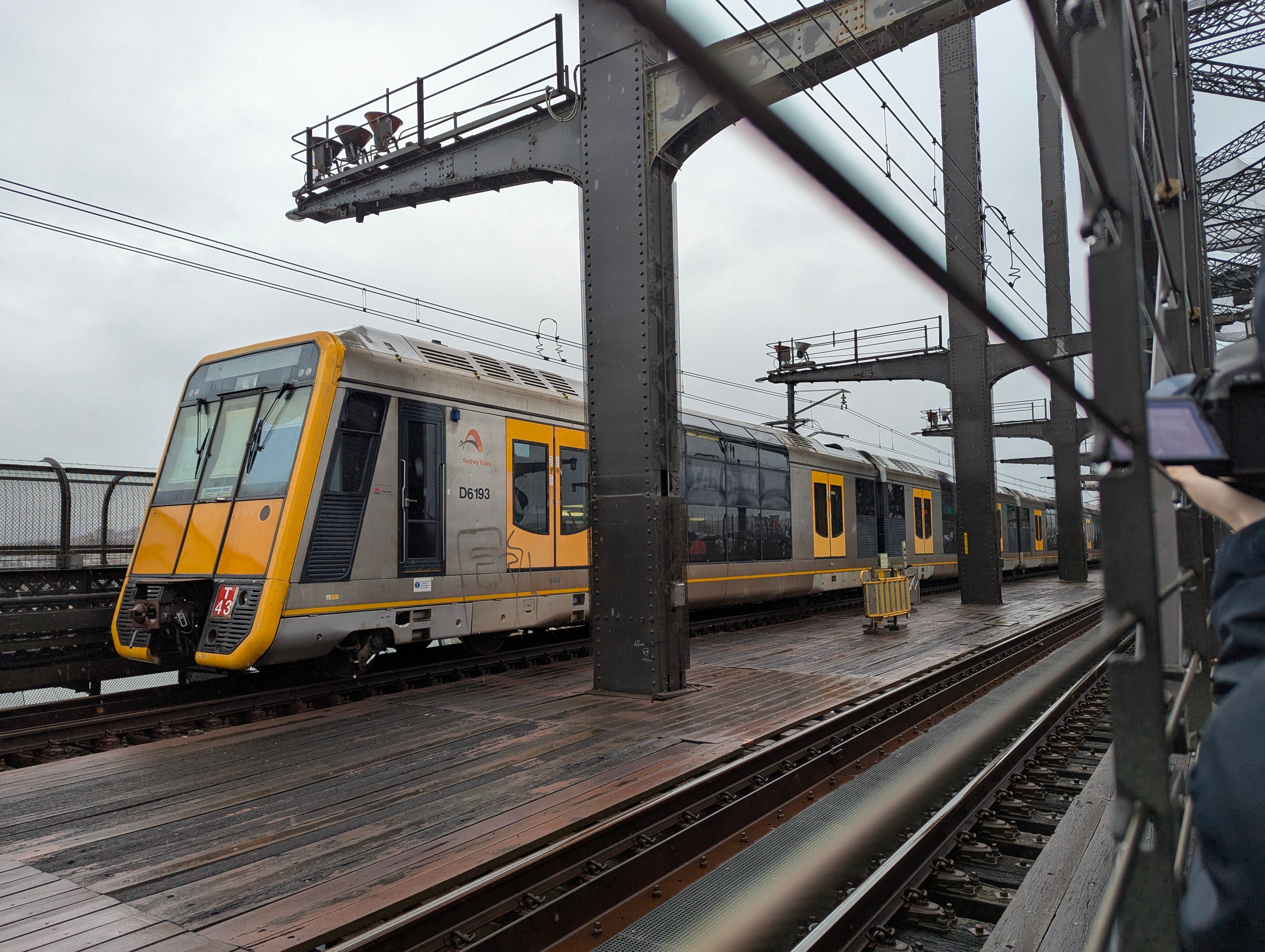 Looking at a double-decker silver "Tangara" suburban train with a distinct slanted yellow front and yellow doors. A fence is visible in the foreground and grey steel bridge pieces are visible at regular intervals to the right of the train. The surfaces around the train are wet and the sky is a dull white colour.
