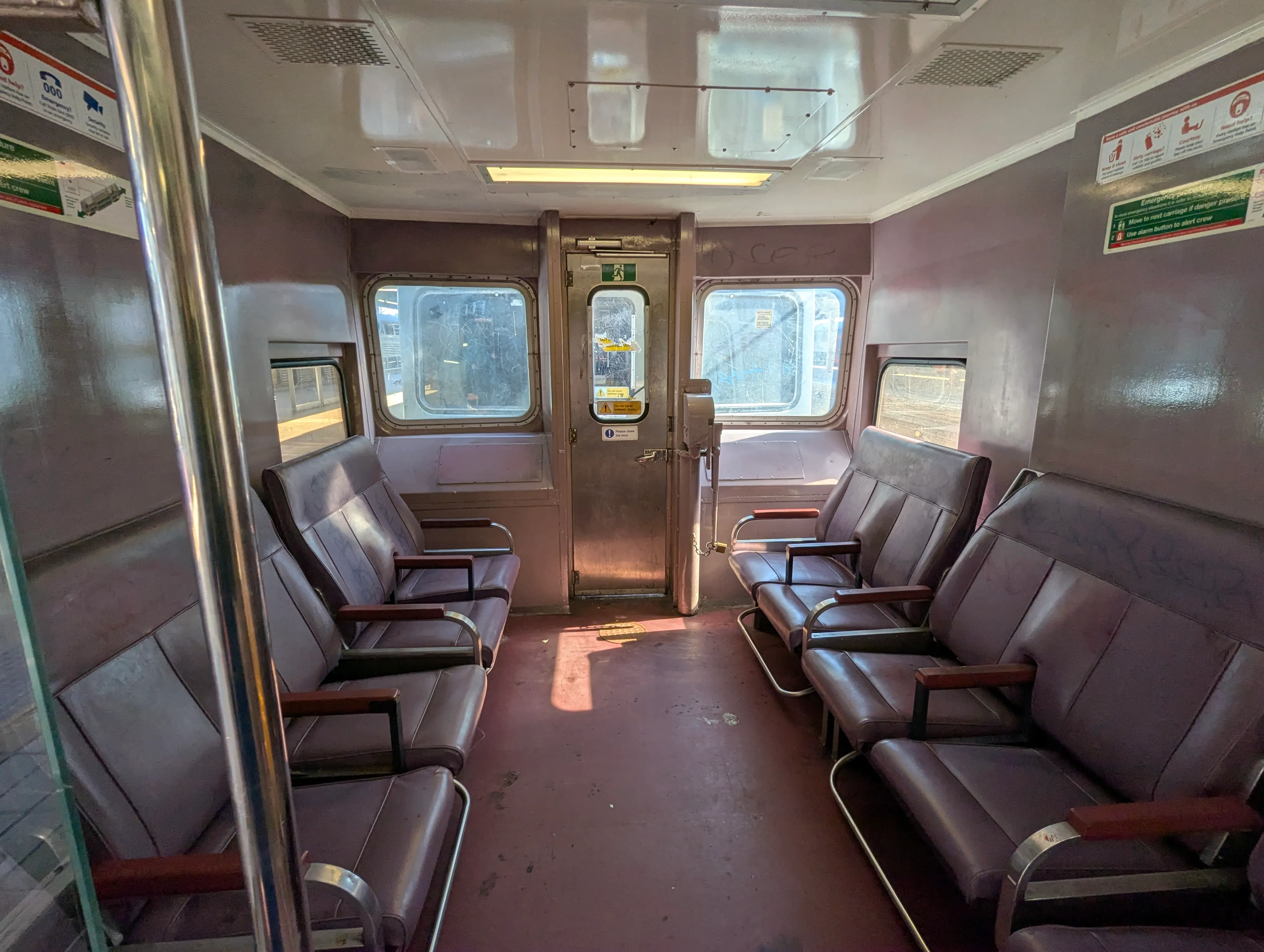 Looking into the vestible area of a converted driving trailer of a V-set, the  purple interior is brightly lit by sunshine and four bush plum colorued seats face inward, two on each side forming a longitudinal seating arrangement. Some faint marks of cleaned graffiti are visible on the chairs. 
