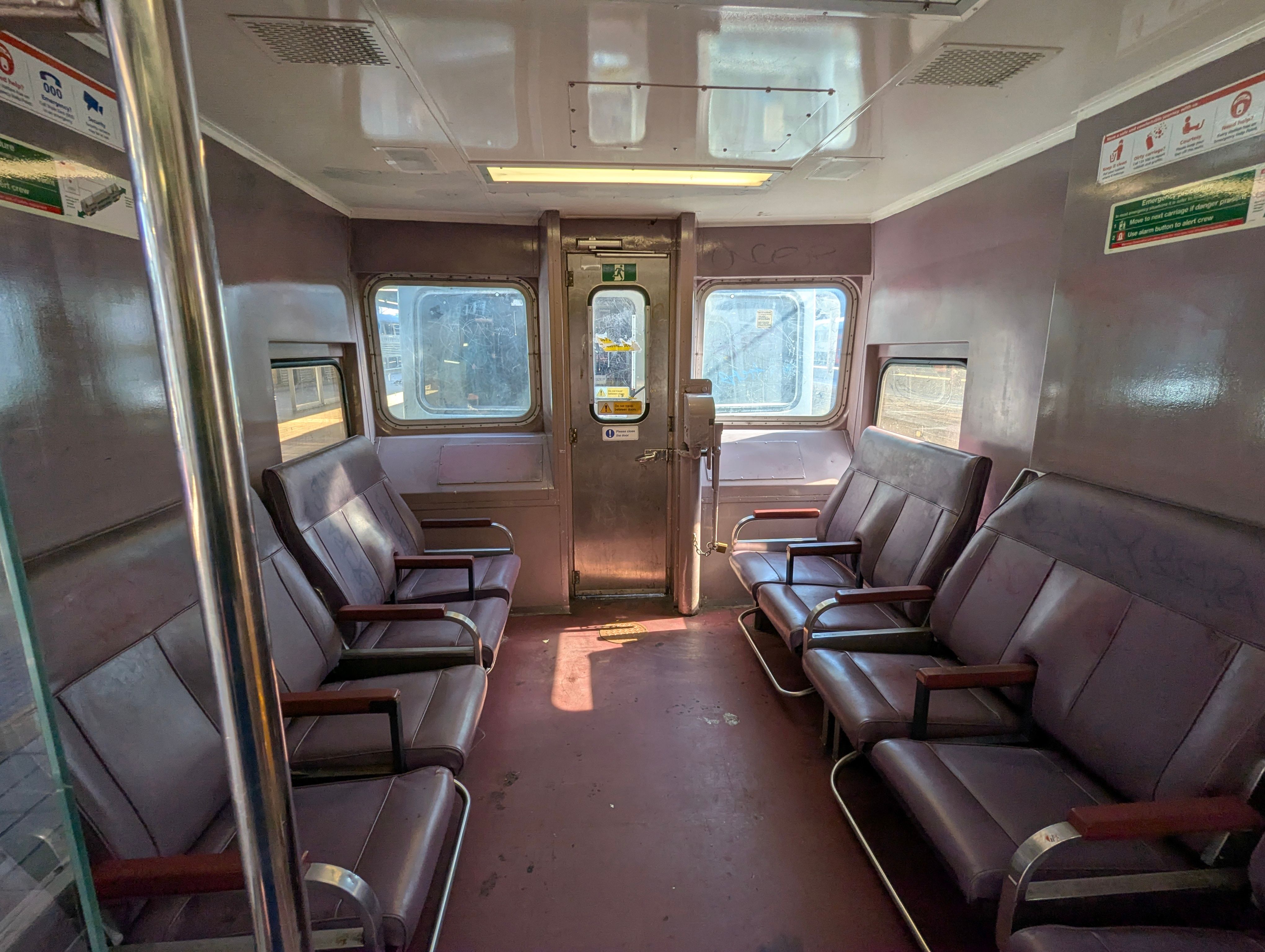 Looking into the vestible area of a converted driving trailer of a V-set, the purple interior is brightly lit by sunshine and four bush plum colorued seats face inward, two on each side forming a longitudinal seating arrangement. Some faint marks of cleaned graffiti are visible on the chairs.