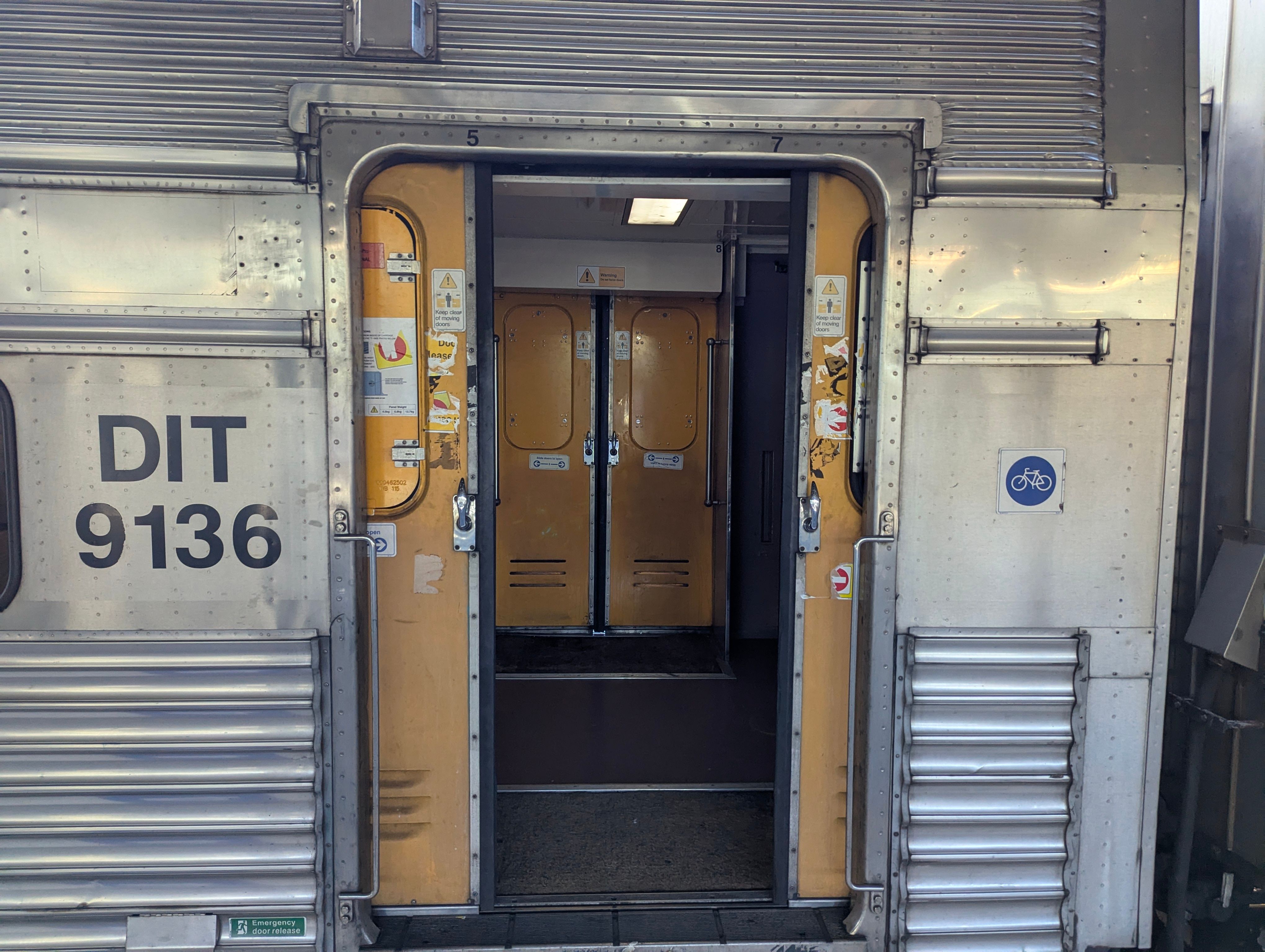 The windows in the external doors of V set carriage DIT 9136 have all been vandalised except one. The doors closest to the camera are ajar, showing the other set of doors having their windows replaced with a temporary steel cover.