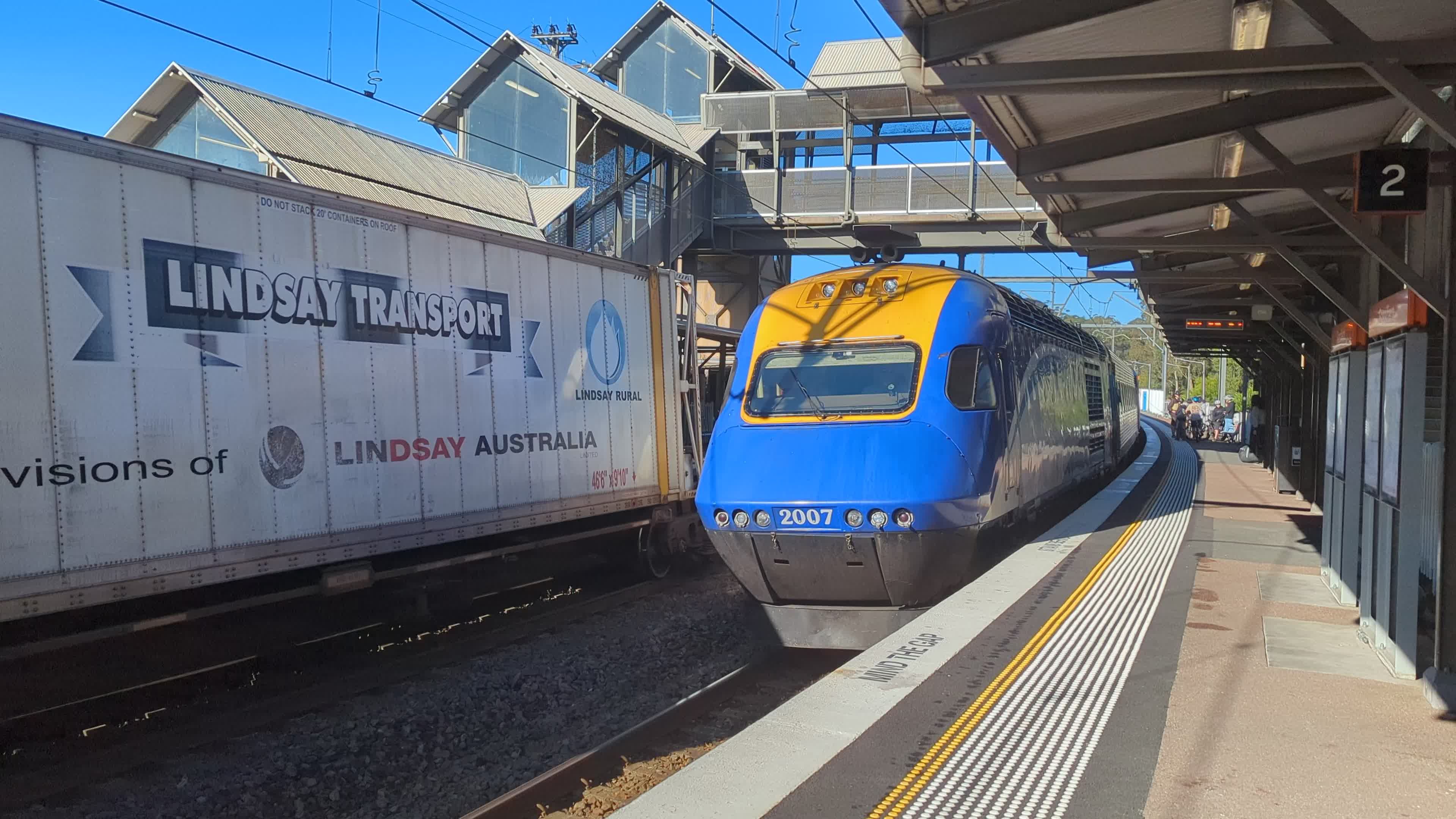 A blue and yellow XPT power car numbered 2007 pulls into the platform with a moving freight train visible on the other track. The overpass of the station is visible in the background and the sky is bright & clear.
