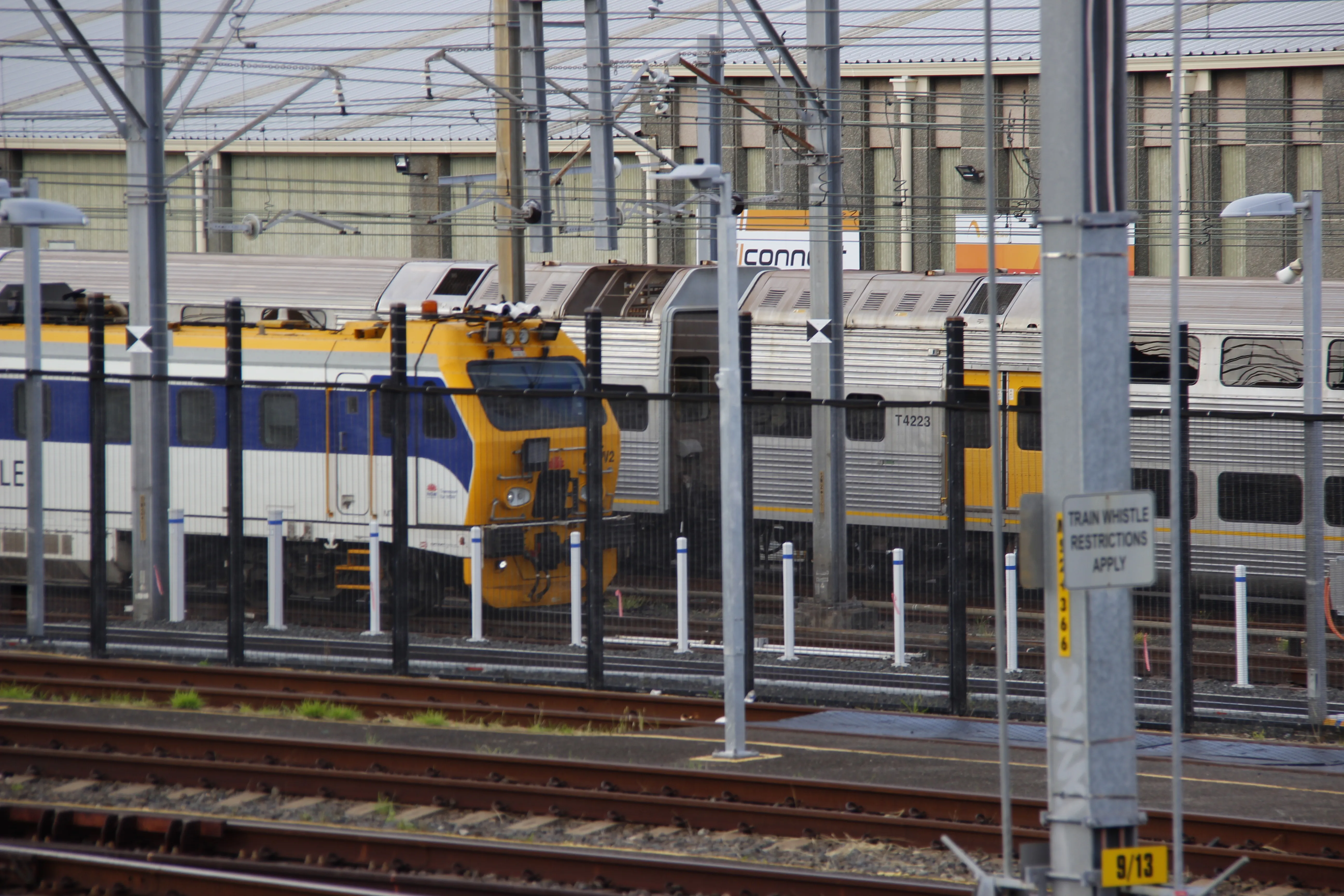 A white, blue and yellow track inspection vehicle (TIV) sitting behind a fence, stabled in a workshop. A double-decker steel K-set is visible behind the TIV. The upper half of the frame is filled with various gantries and supports that hold up the overhead wires in the area. A couple pairs of tracks are visible in the lower half of the frame.