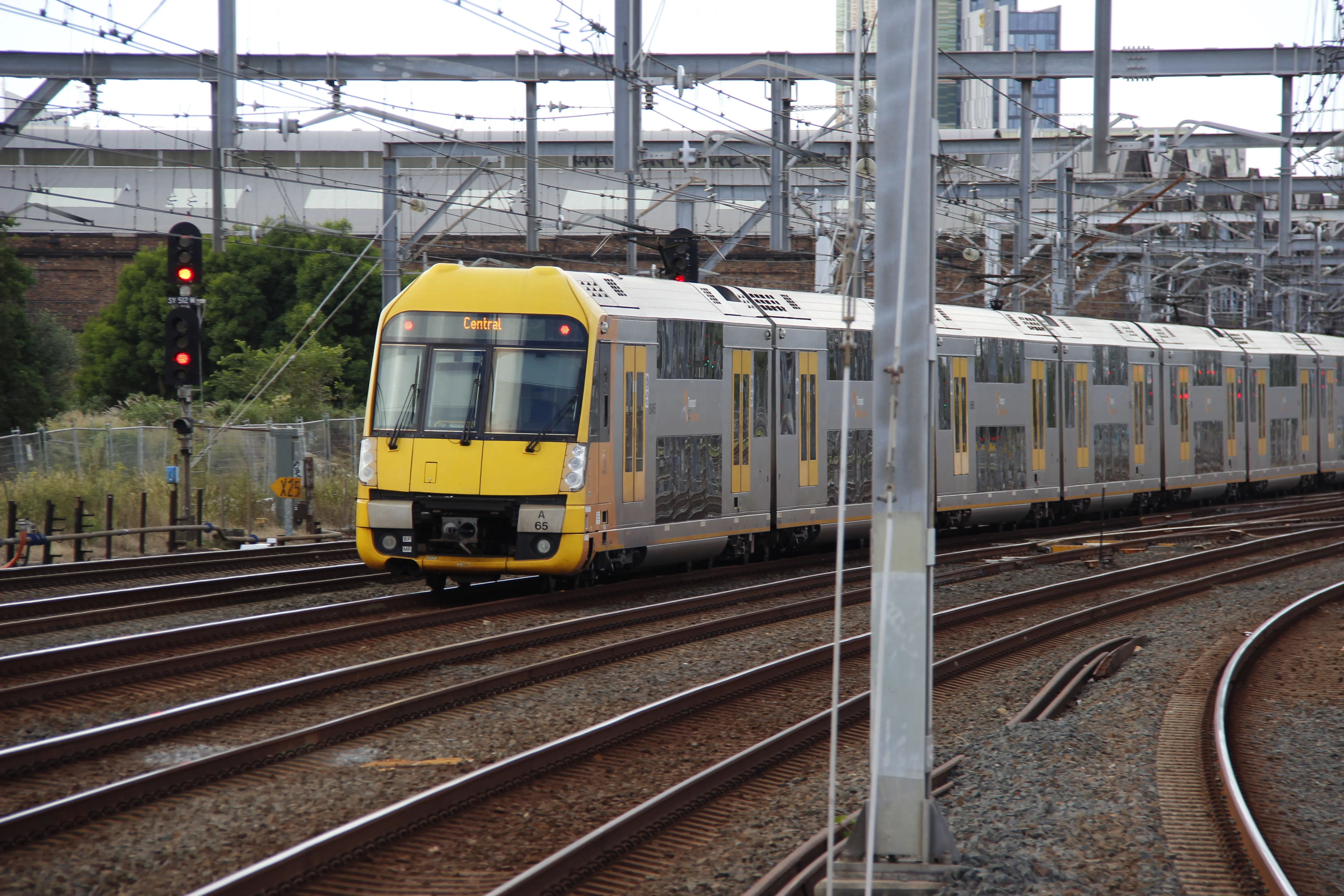A yellow and silver double-decker suburban train labelled "A65" on the third track from the left out of six tracks. Above its windshield, the electronic  destination board reads "Central". Lots of overhead wiring and steel gantries are visible toward the top of the frame. In the background, some brick buildings can be seen. The sky is overcast being an even white colour.