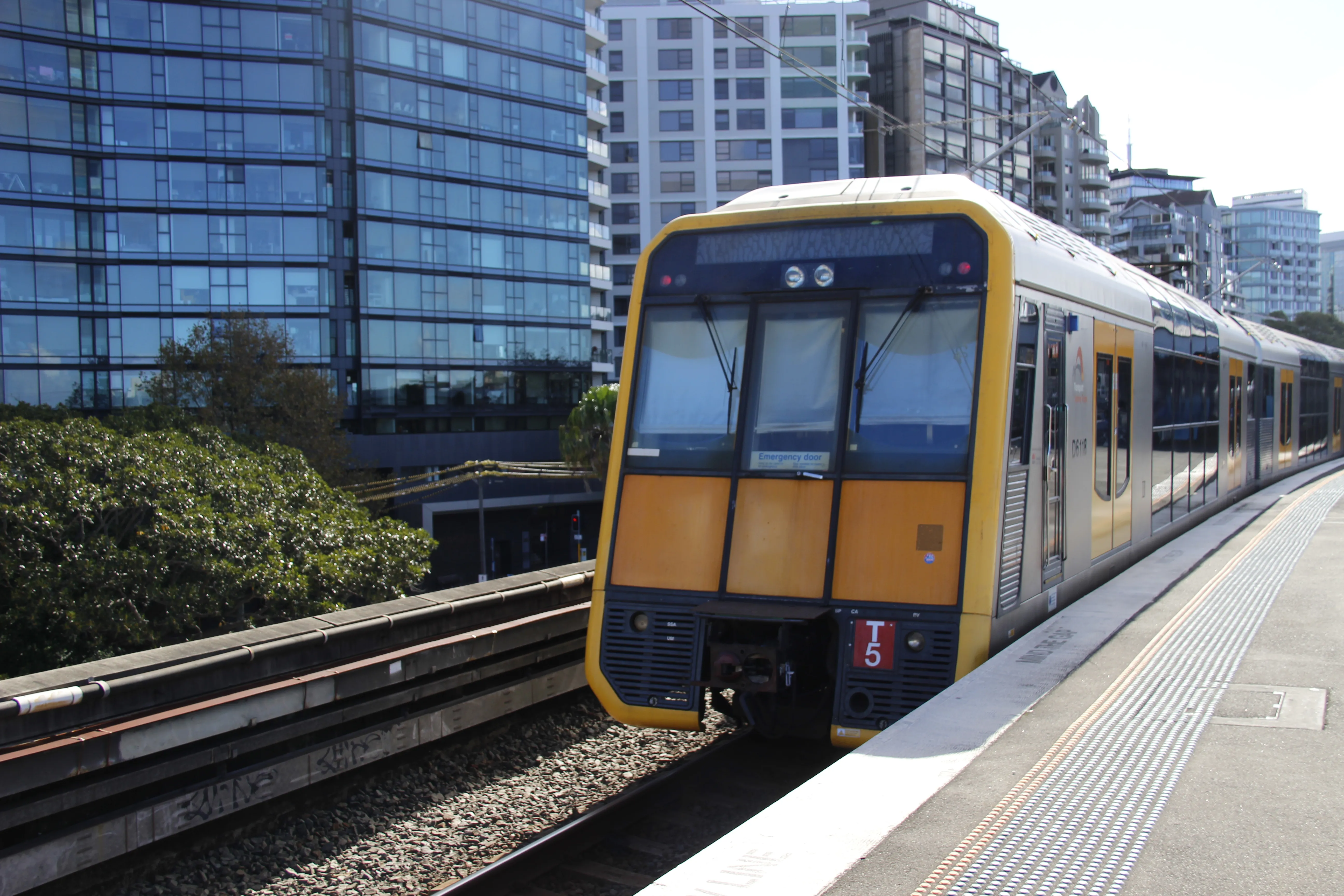 A yellow, black and silver double-decker Tangara train at the platform. The rear of the train is visible, which has a slanted flat design which is yellow and black below the windshield. A red target plate is visible reading "T5". Above the windshield is an empty (probably broken) destination board. Only the rear two carriages are visible in the frame, with yellow doors and a curved window design on the top floor. The scene is brightly lit with ample sunshine and the sky is an overexposed white colour. There are apartment buildings visible in the background.