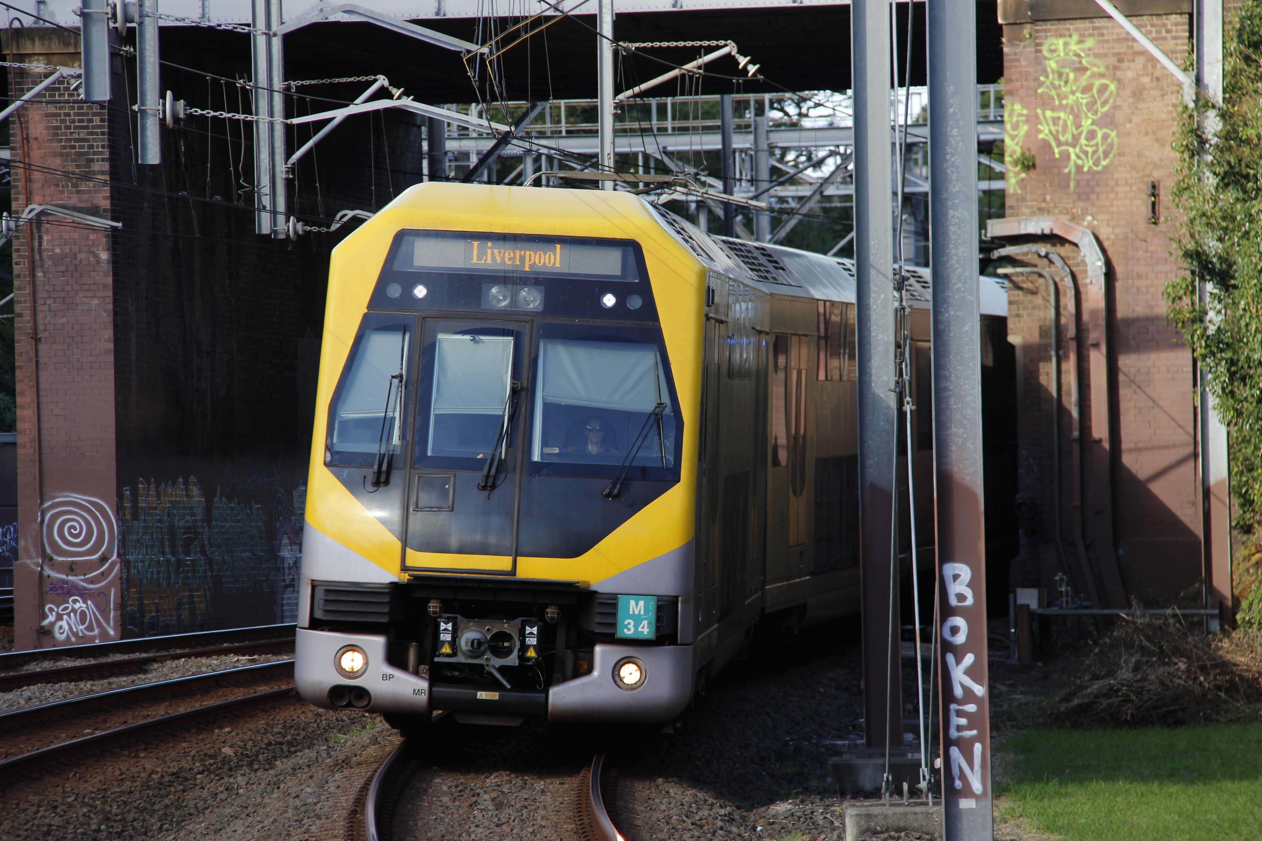 A bright yellow and grey passenger train exiting a brick railway underpass, slightly curving towards the camera. The destination board of the train reads "Liverpool" in a serif font. Overhead electrical wiring and support structures are visible. Some graffiti is present on the underpass' brick walls.