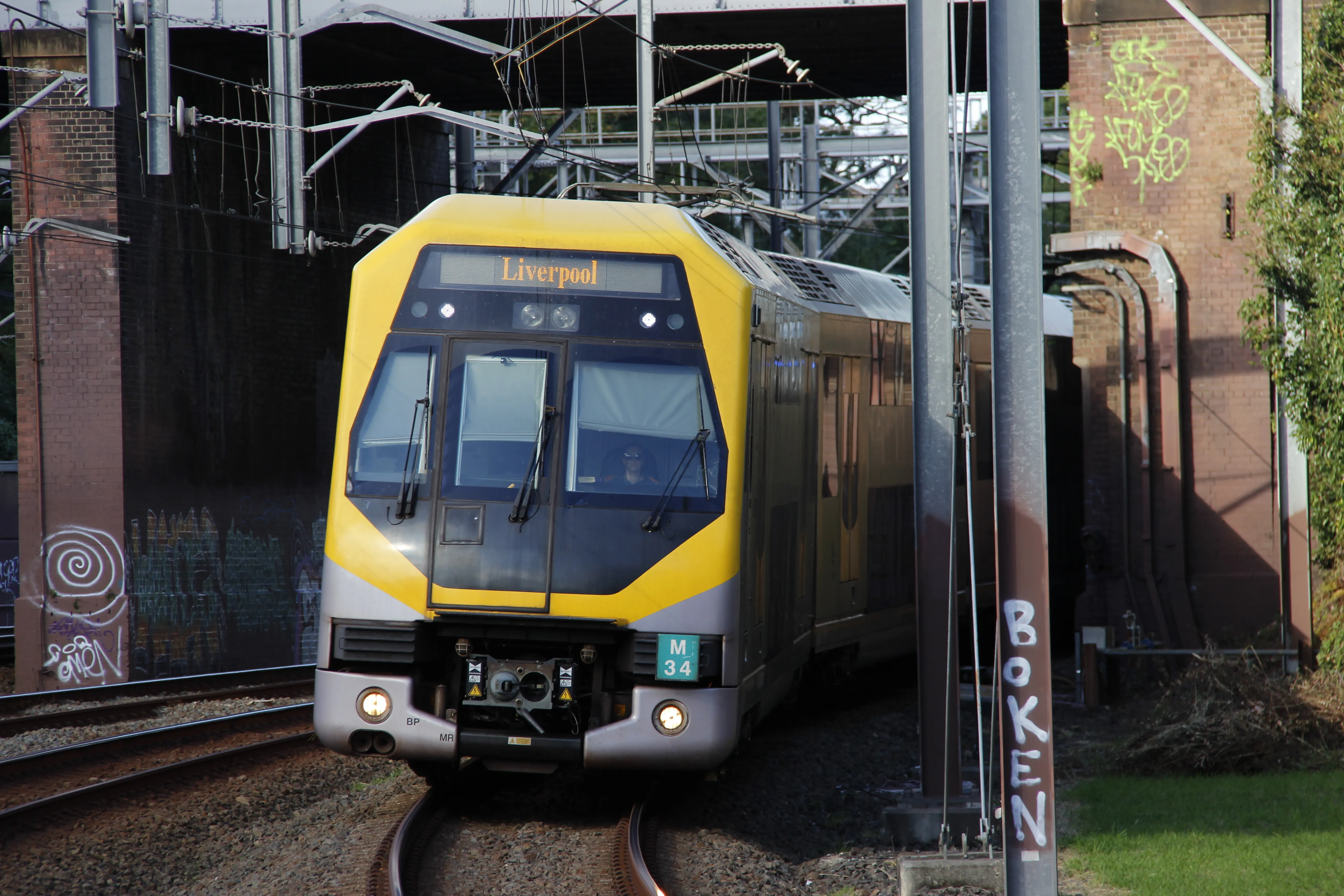 A bright yellow and grey passenger train exiting a brick railway underpass,  slightly curving towards the camera. The destination board of the train reads "Liverpool" in a serif font. Overhead electrical wiring and support structures are visible. Some graffiti is present on the underpass' brick  walls.