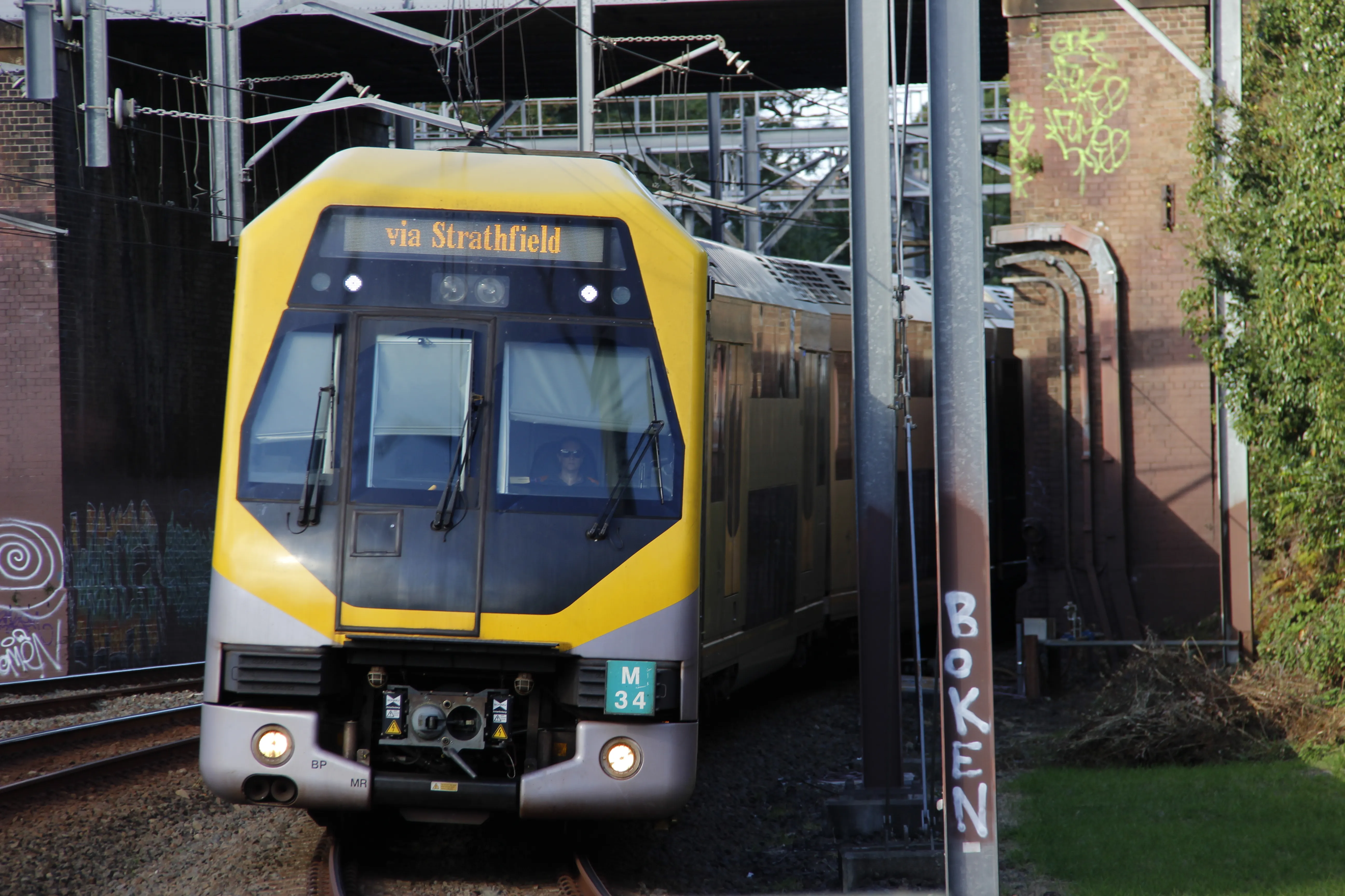 A bright yellow and grey passenger train exiting a brick railway underpass,  slightly curving towards the camera. The destination board of the train reads "via Strathfield" in a serif font. Overhead electrical wiring and support structures are visible. Some graffiti is present on the underpass' brick  walls.