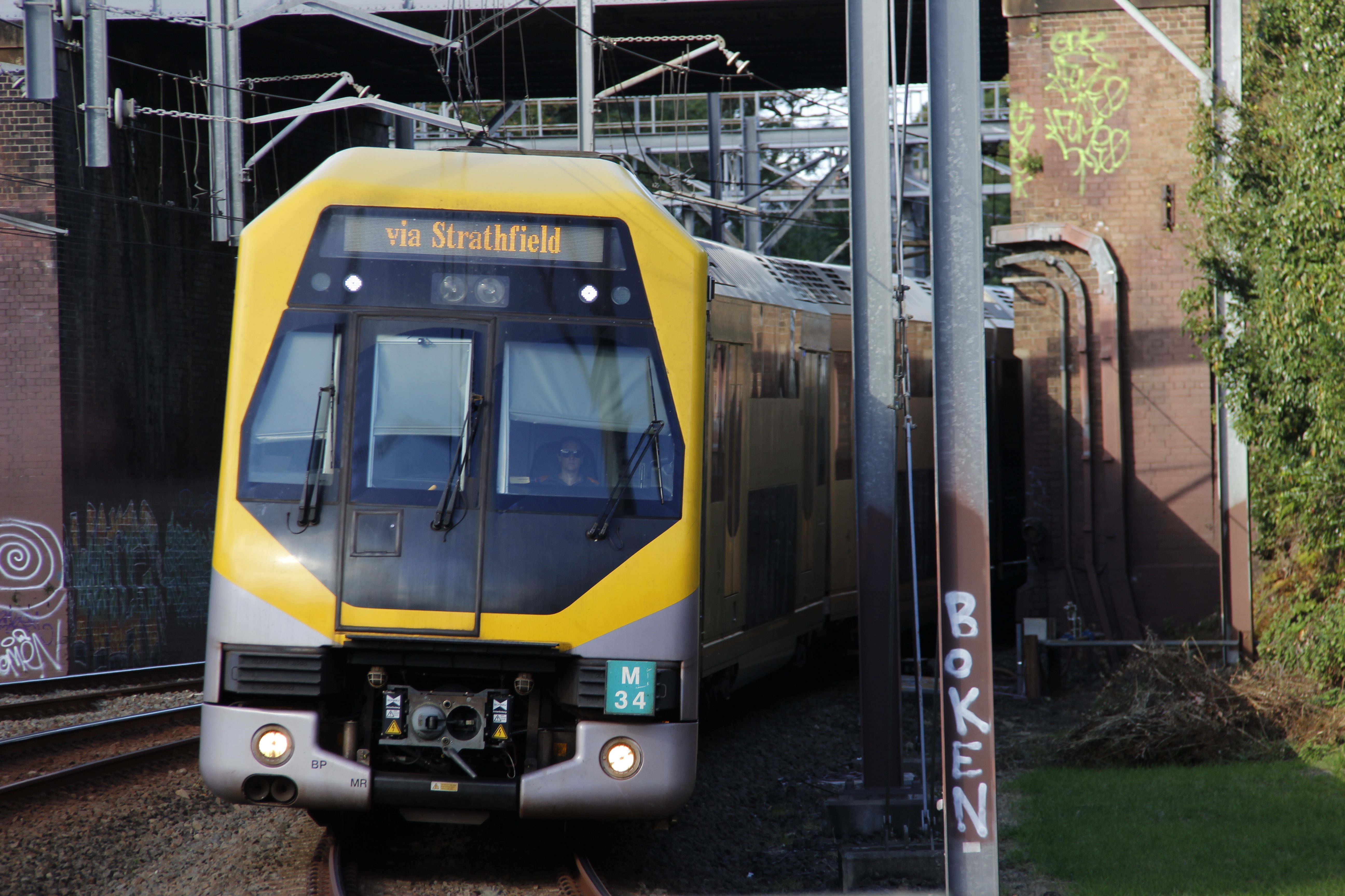 A bright yellow and grey passenger train exiting a brick railway underpass, slightly curving towards the camera. The destination board of the train reads "via Strathfield" in a serif font. Overhead electrical wiring and support structures are visible. Some graffiti is present on the underpass' brick walls.