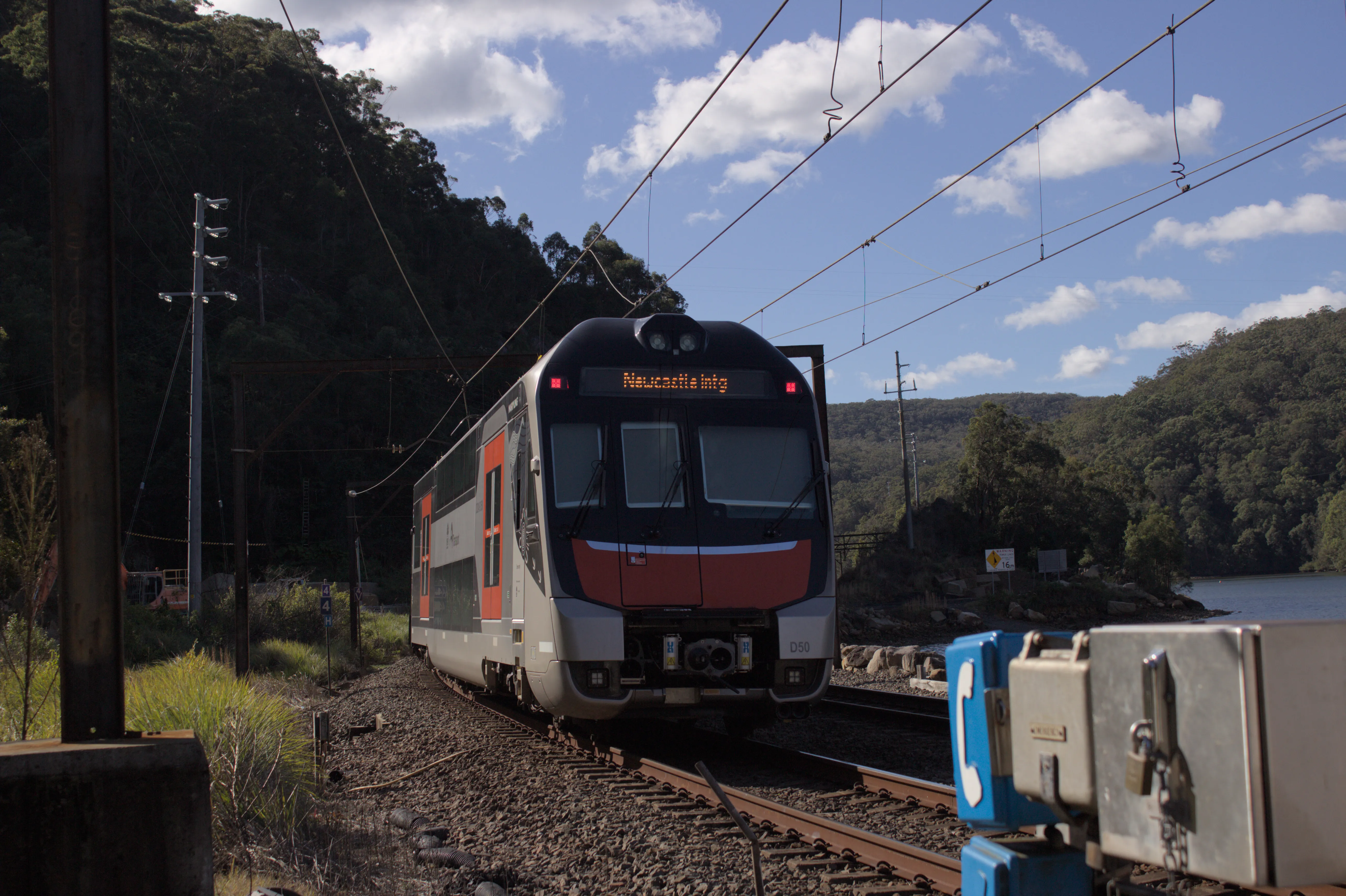 A modern Mariyung double-decker intercity train travelling away from the camera with the rear of the train visible, along curved tracks on top of a causeway. There is a pair of tracks, and the train is travelling on the left track. The train is mostly silver with orange-accented doors and black highlights. The electronic destination board is labelled "Newcastle Intg". Overhead wires are visible with a rusty supporting pole out of focus to the left of the frame. In the foreground, a bright blue telephone box and some locked steel boxes are visible. The background has green forested hills and a body of water  (Mullet Creek) is visible to the right of the causeway. The sky is clear with  scattered clouds and it is broad daylight. 