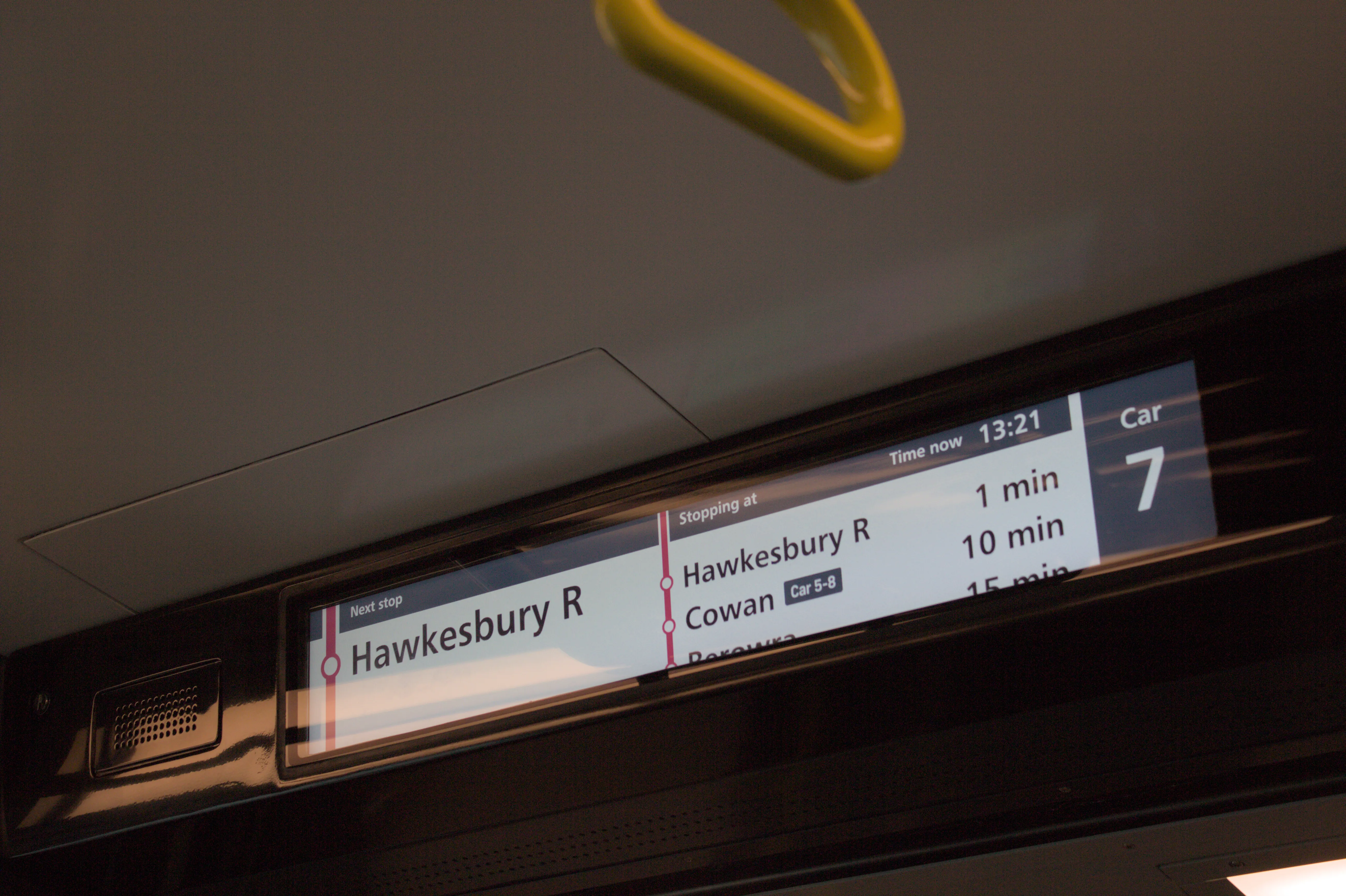 An electronic passenger information screen mounted near the ceiling, bordered in black. Displayed on the screen is stop information, showing that the next stop is "Hawkesbury R", and that the proceeding stations are Cowan (only accessible from Cars 5-8) in 10 minutes and Berowra in 15 minutes. Above the list of stations the current time is shown as 13:21 and on the far right of the screen the car number is listed in as "Car 7". The ceiling panels are a clean white colour  and an out of focus bright yellow grab handle is visible to the top of the  frame. The overall lighting is warm.