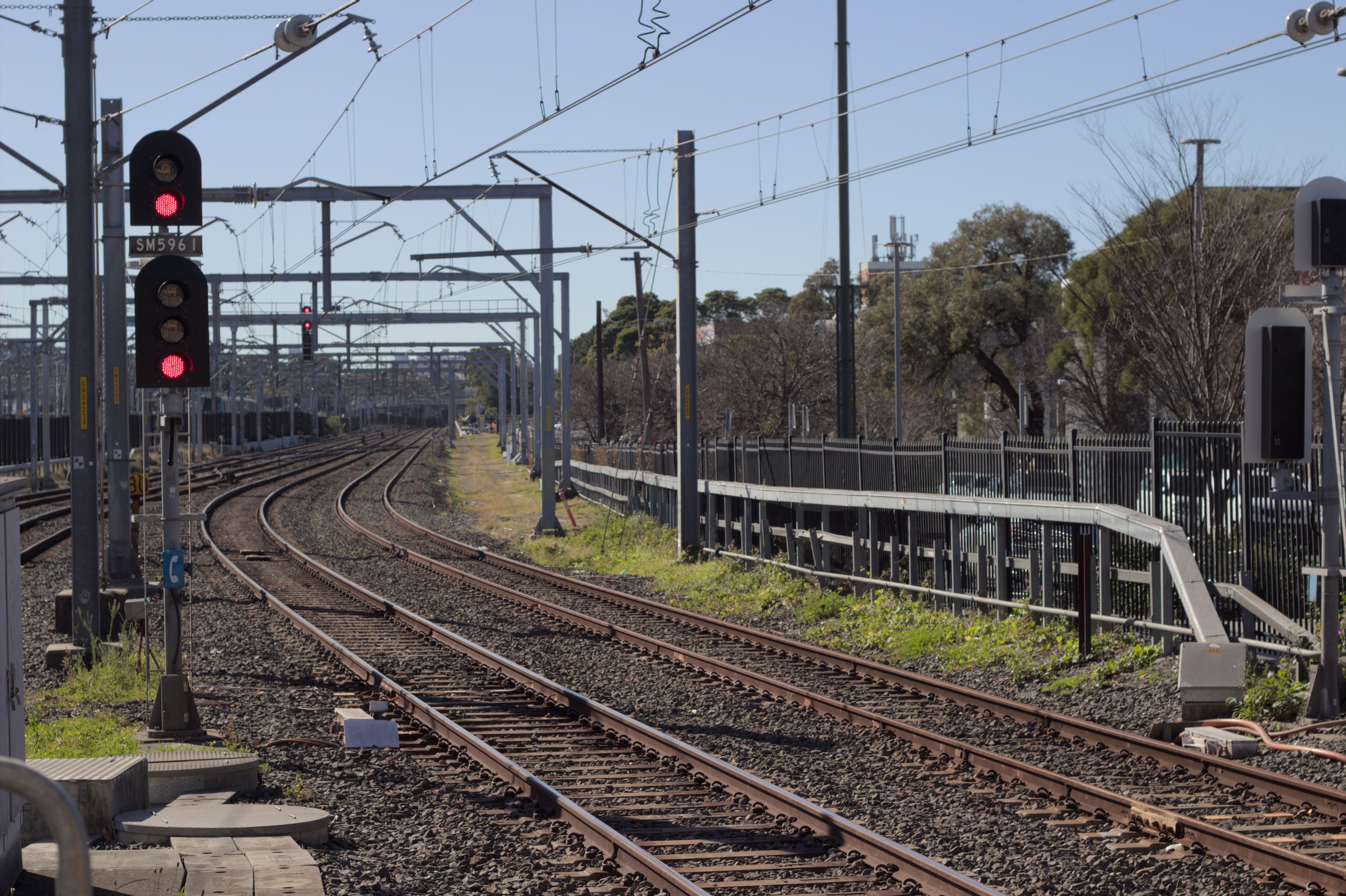 Looking into the rail corridor from the end of the platform, a signal can be seen prominently to the left of the frame, displaying two red aspects with a small board reading "SM5961" in between the two signals on the pole. Two tracks are immediately to the right side of the signal toward the bottom centre of the frame, with a pair of offset signals facing the other direction visible on the far right of the image, for the right track. Two more tracks can be seen merging into the rail corridor further ahead, and overhead wires and gantries fill the top of the image.