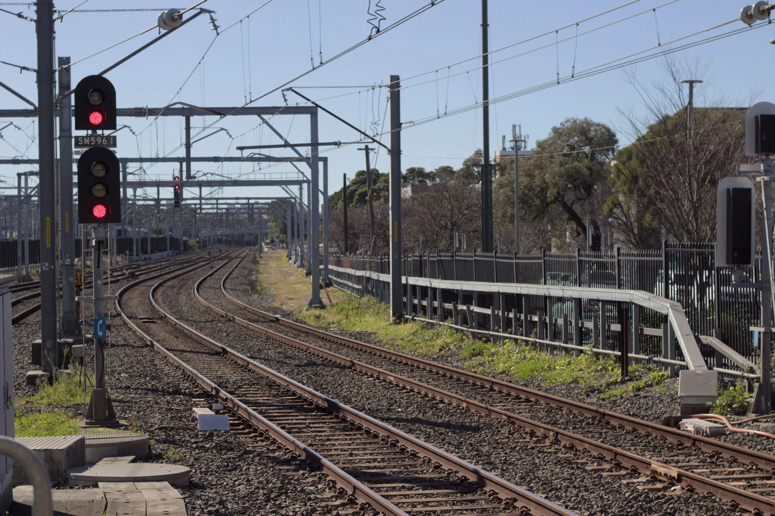 Looking into the rail corridor from the end of the platform, a signal can be  seen prominently to the left of the frame, displaying two red aspects with a  small board reading "SM5961" in between the two signals on the pole. Two tracks are immediately to the right side of the signal toward the bottom centre of the frame, with a pair of offset signals facing the other direction visible on the far right of the image, for the right track. Two more tracks can be seen merging into the rail corridor further ahead, and overhead wires and gantries fill the  top of the image. 