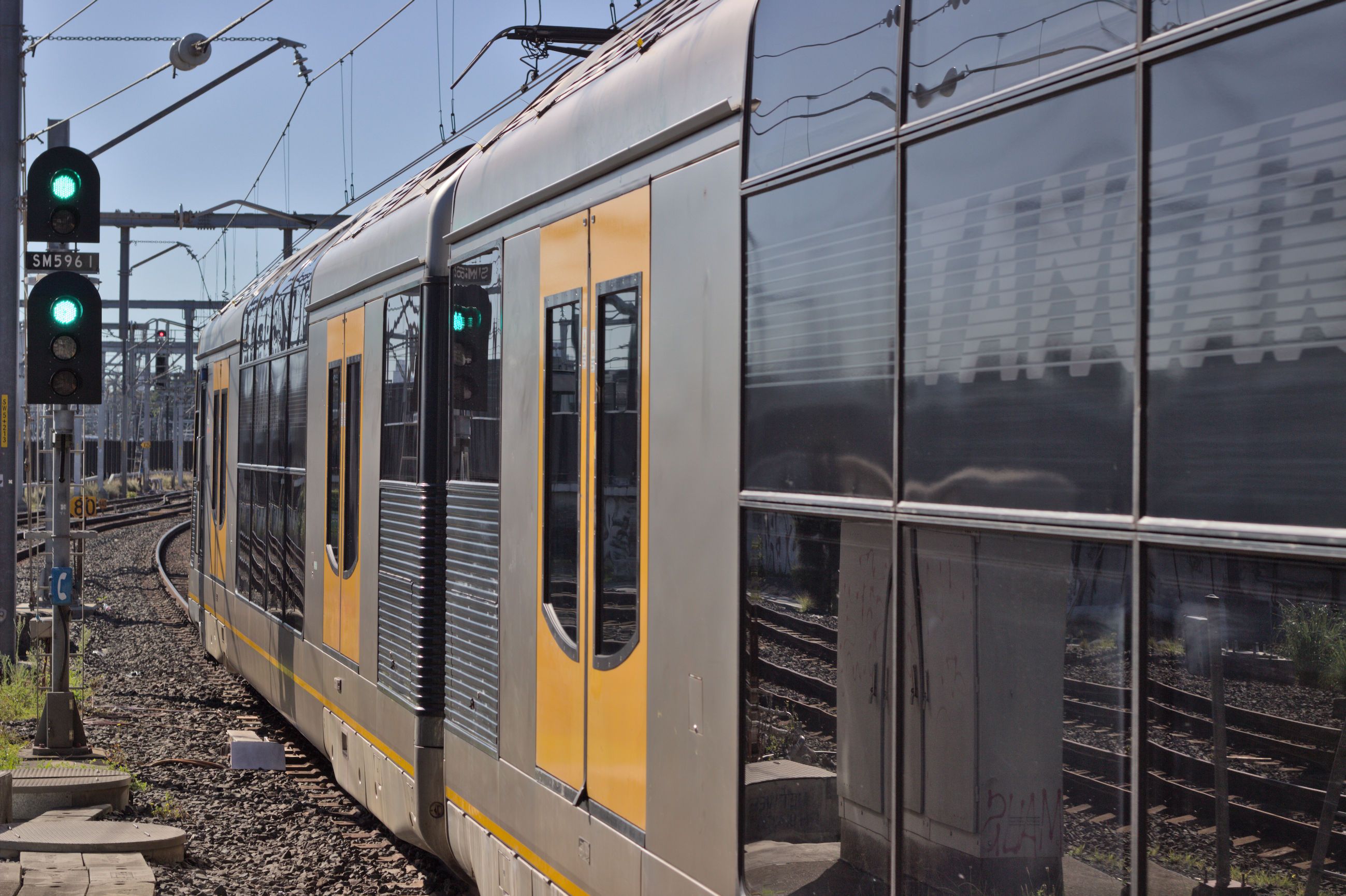 A double-decker suburban electric "Tangara" train is seen departing, passing a prominent green on green (double green) signal to the left of the frame. The doors of this train are bright yellow with the body being a stainless steel silver and the windows being a reflective black, whilst the upper floor windows curve slightly. Only the first carriage and half of the second carriage are visible. The shot was taken looking out at the end of the platform into the rail corridor but no parts of the platform itself are actually visible. Overhead wires and some supporting equipment are visible toward the top of the frame along with the pantograph attached to the end of the first carriage.