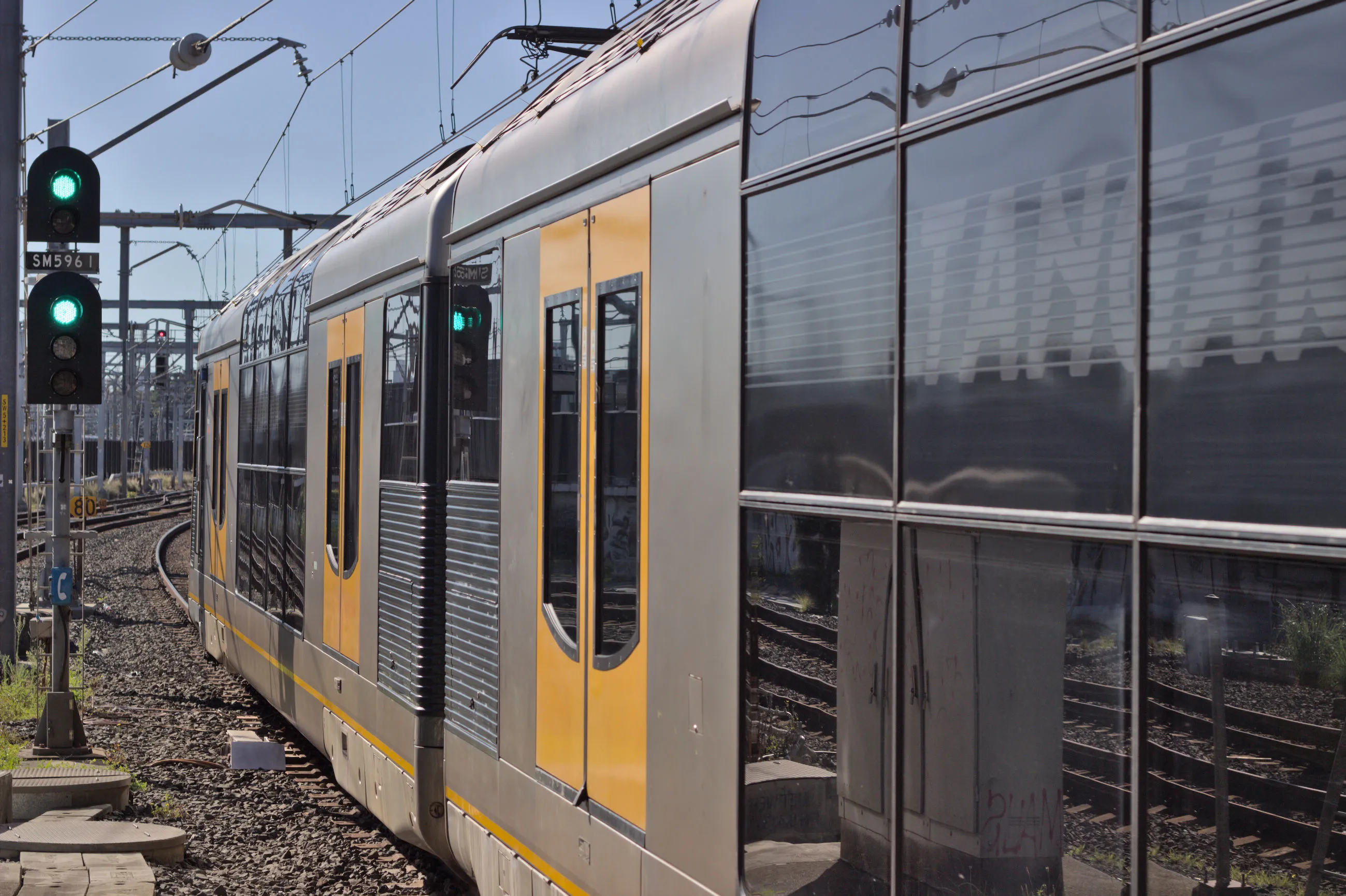 A double-decker suburban electric "Tangara" train is seen departing, passing a prominent green on green (double green) signal to the left of the frame. The  doors of this train are bright yellow with the body being a stainless steel silver and the windows being a reflective black, whilst the upper floor windows curve slightly. Only the first carriage and half of the second carriage are visible. The shot was taken looking out at the end of the platform into the  rail corridor but no parts of the platform itself are actually visible.  Overhead wires and some supporting equipment are visible toward the top of the frame along with the pantograph attached to the end of the first carriage.