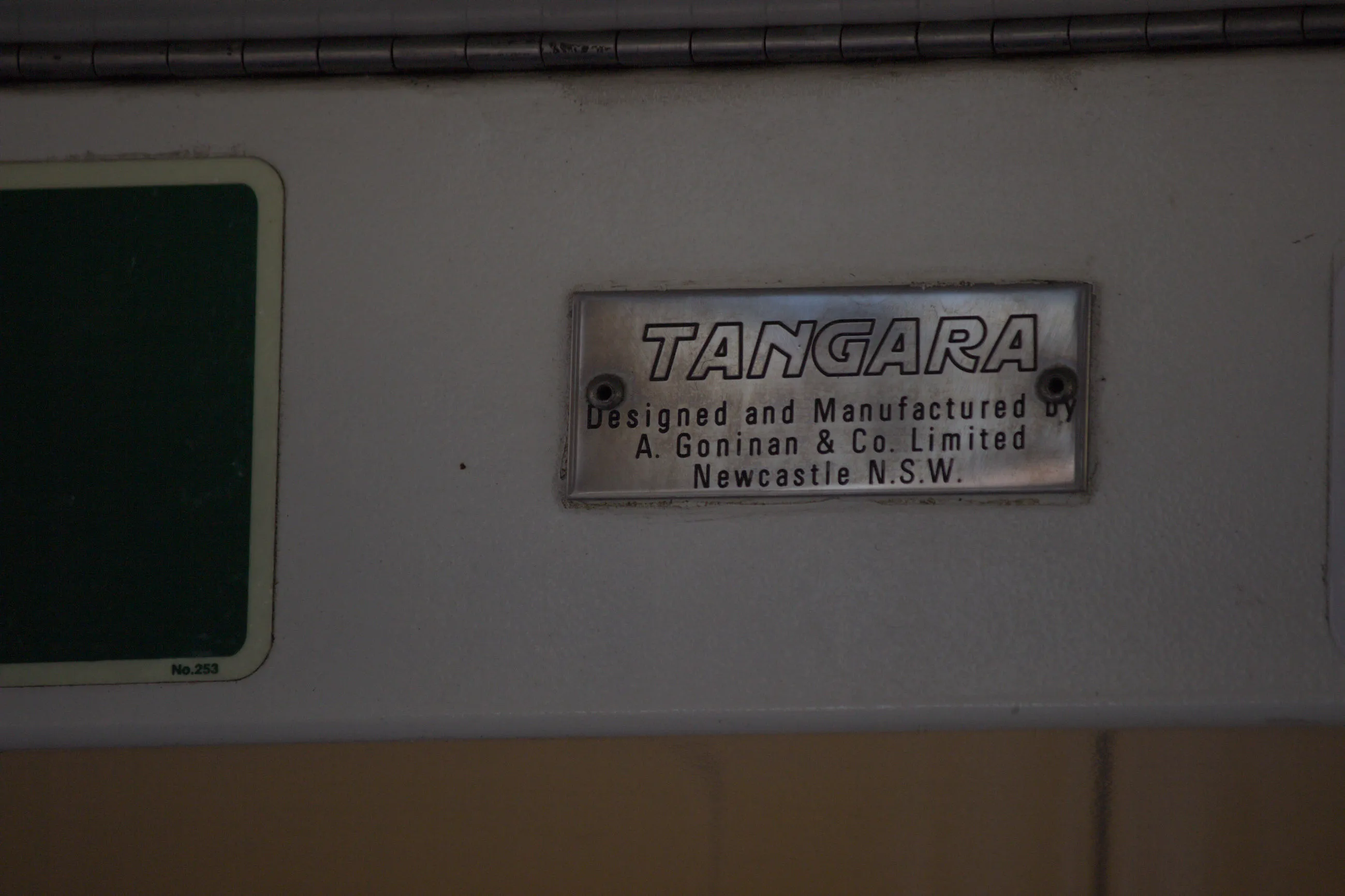A small shiny rectangular steel plate reading "TANGARA" in uppercase writing, and reading "Designed and Manufactured by A. Goninan & Co. Limited Newcastle N.S.W." under it in smaller print. The plate is held into place with two  bolts at the centre of each side. The background is a clear white colour and  the image appears a bit dark. A bit of a green exit sticker is visible to the  left of the frame but is cut off.