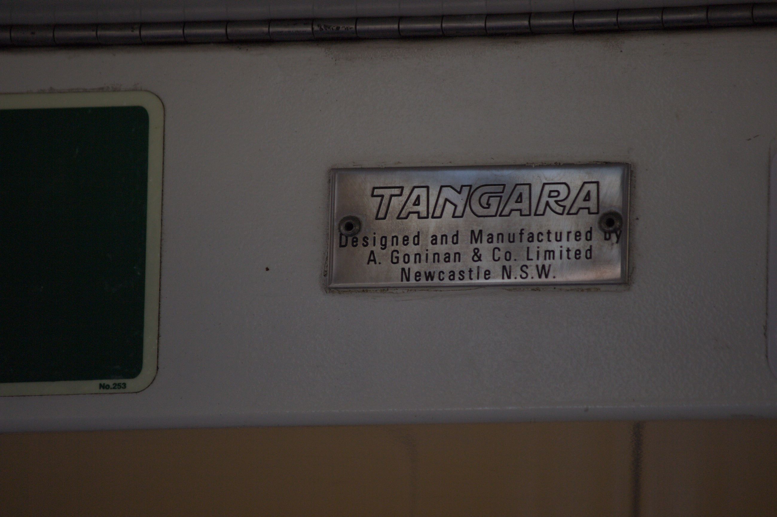 A small shiny rectangular steel plate reading "TANGARA" in uppercase writing, and reading "Designed and Manufactured by A. Goninan & Co. Limited Newcastle N.S.W." under it in smaller print. The plate is held into place with two bolts at the centre of each side. The background is a clear white colour and the image appears a bit dark. A bit of a green exit sticker is visible to the left of the frame but is cut off.