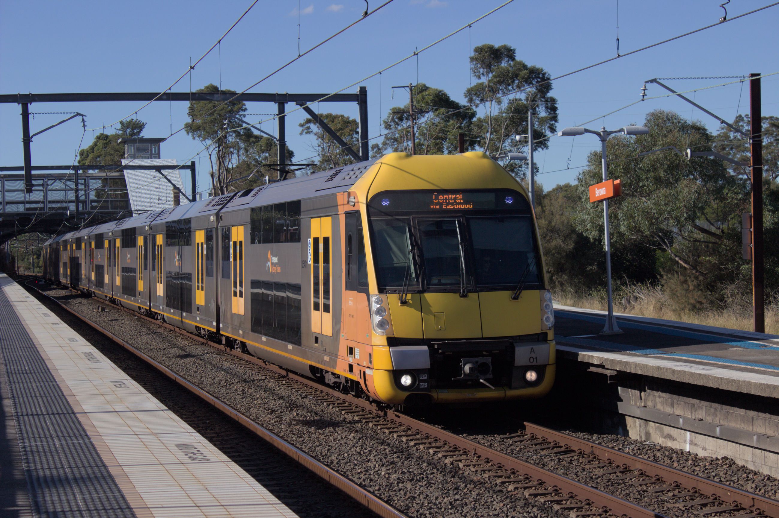 A modern double-decker suburban train with a yellow front and a silver body, with yellow highlights around the doors and dark windows at a platform. The destination board on the front above the windshield reads "Central via Eastwood". All 8 carriages of the train are visible. Another platform which the photographer is standing on is visible in the shot as well. In the background there is a footbridge over the tracks and some trees. Overhead wiring is visible in the top of the frame as well as a wire gantry toeard the middle of the platform supporting these wires. THe sky is a blue colour and it is bright daylight.