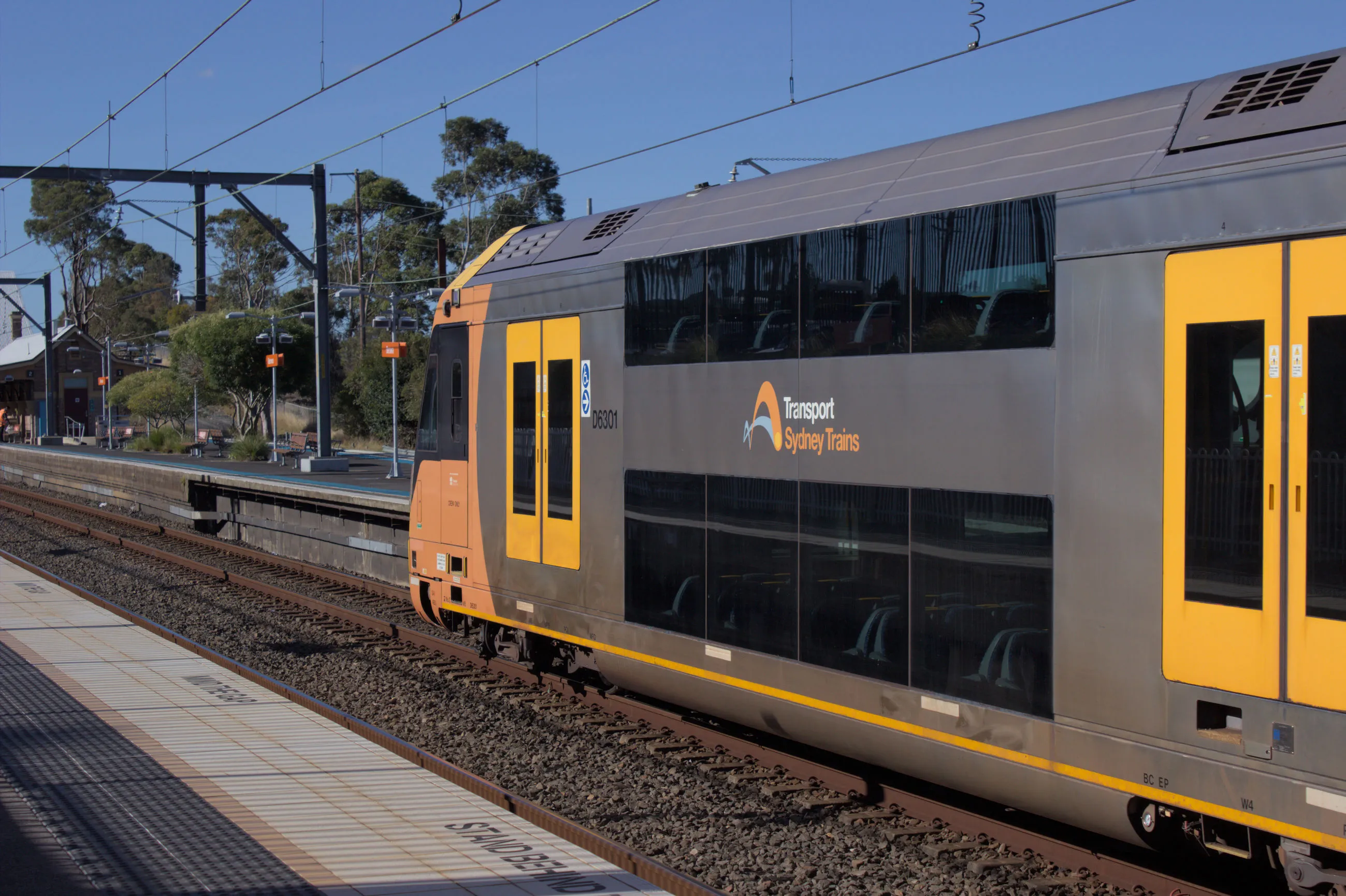 A modern double-decker suburban train departing from a platform. The train has a silver body and yellow doors