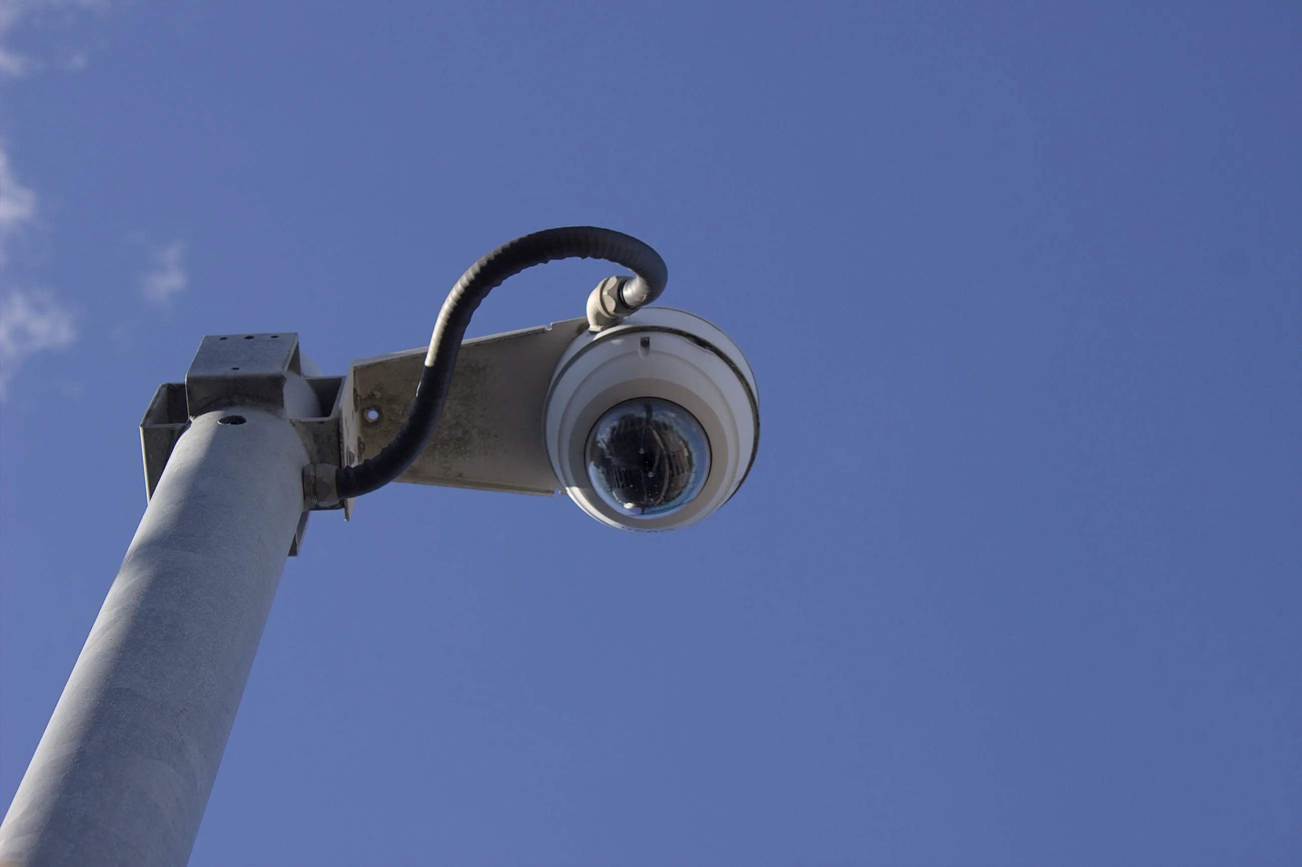 A metal pole supporting a white round security camera can be seen, with the  security camera mounted to the right of the pole so it occupies the centre of the frame. The photo is taken from the ground looking upward so only the top of the pole, the camera and the sky are visible. A thick black wire runs from the side of the camera to the pole. The sky is a mostly clear blue colour with some wispy clouds visible to the far left of the frame. 