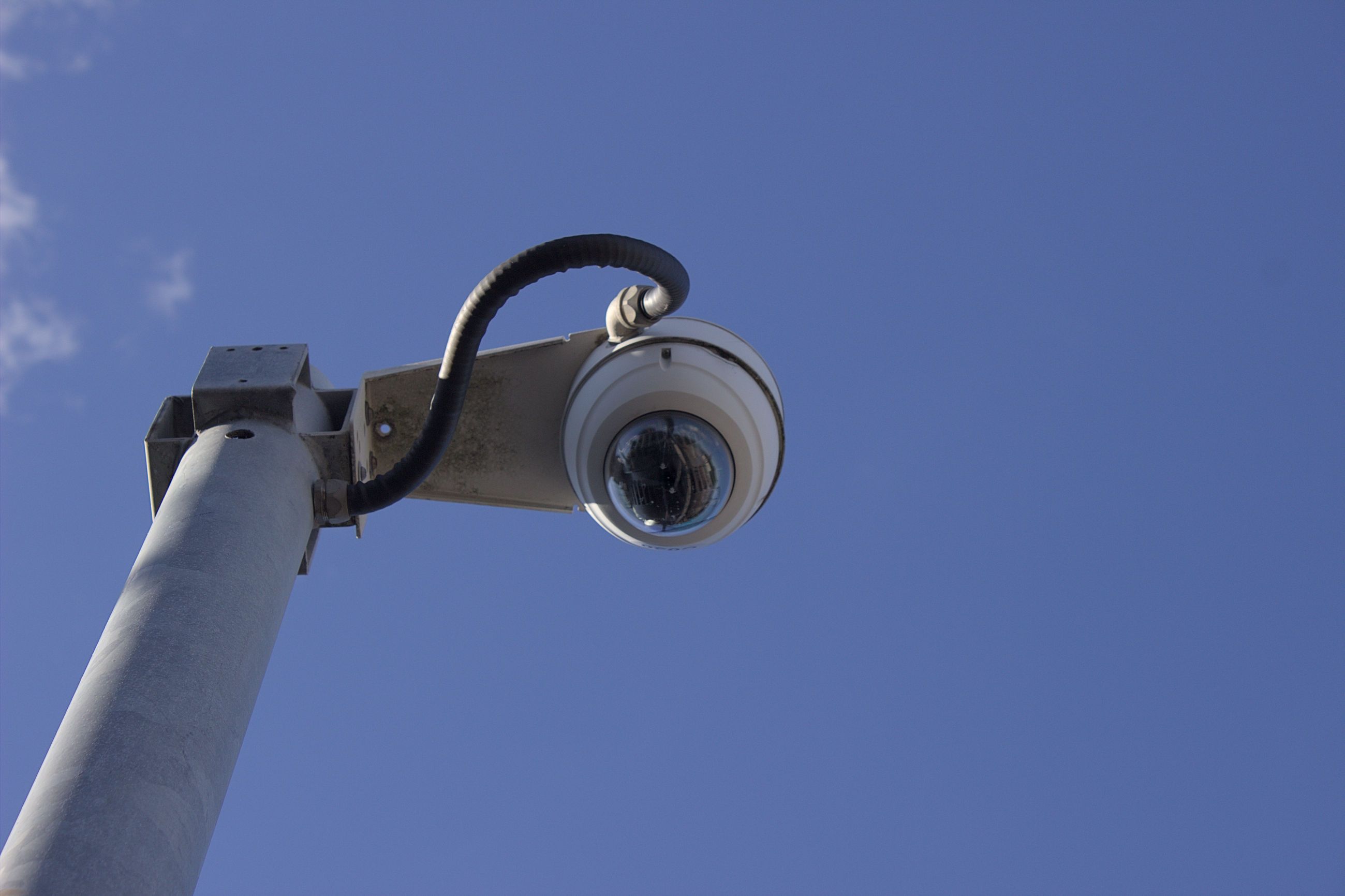 A metal pole supporting a white round security camera can be seen, with the security camera mounted to the right of the pole so it occupies the centre of the frame. The photo is taken from the ground looking upward so only the top of the pole, the camera and the sky are visible. A thick black wire runs from the side of the camera to the pole. The sky is a mostly clear blue colour with some wispy clouds visible to the far left of the frame.
