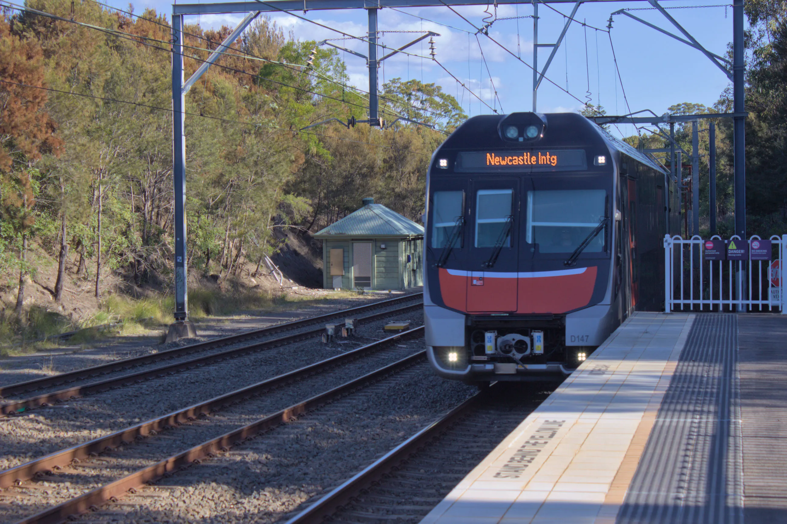A modern double-decker train passing the platform threshold at a semi-rural  station surrounded by trees. the train's front is primarily black with silver bumpers and a red accent below the three-section windshield, and an electronic destination display reading "Newcastle Intg" above the windshield. A platform can be seen to the right of the train with some tactile paving and safety  lettering in black reading "Stand Behind the Yellow Line", out of focus. To  the left of the train are two more tracks and there are overhead wires supported by a gantry just beyond the platform's end.