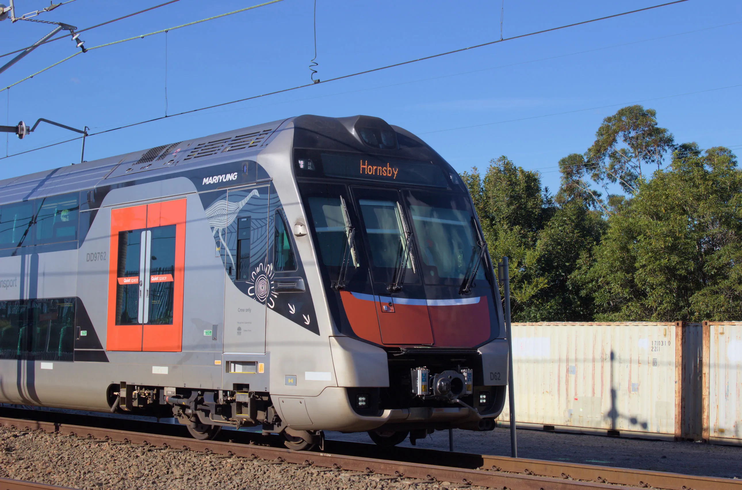 A double-decker Mariyung train travelling along a railway line with trees and two white shipping containers in the background. The train is viewed side on with only the front and first door visible, featuring a prominent curved design and a silver body with red-orange accents on the doors and front. The destination board above the windscreen on the front of the train reads 'Hornsby'. Overhead wires are visible to the top of the frame, and it is a clear sunny day.