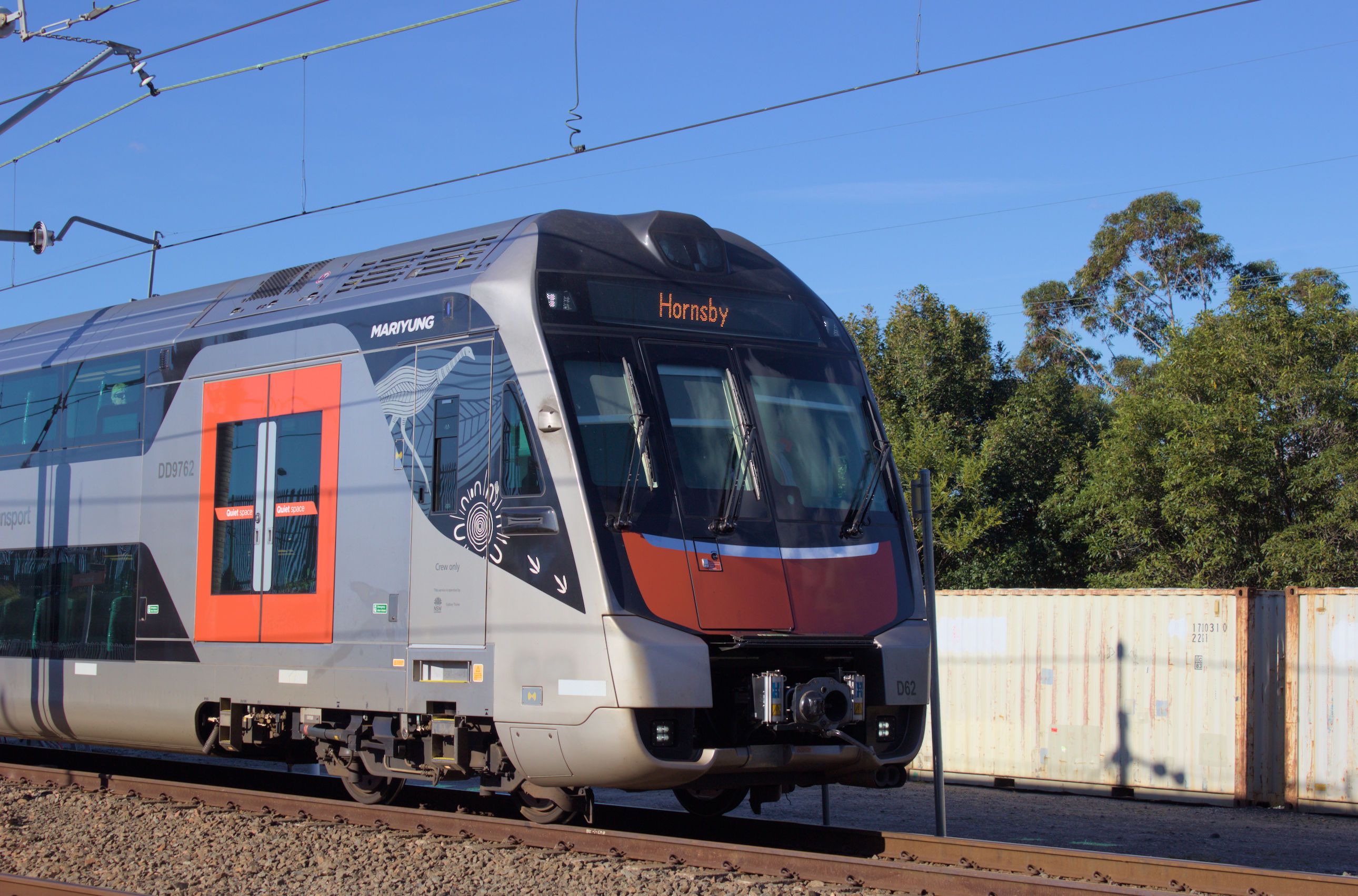A double-decker Mariyung train travelling along a railway line with trees and two white shipping containers in the background. The train is viewed side on with only the front and first door visible, featuring a prominent curved design and a silver body with red-orange accents on the doors and front. The destination board above the windscreen on the front of the train reads 'Hornsby'. Overhead wires are visible to the top of the frame, and it is a clear sunny day.