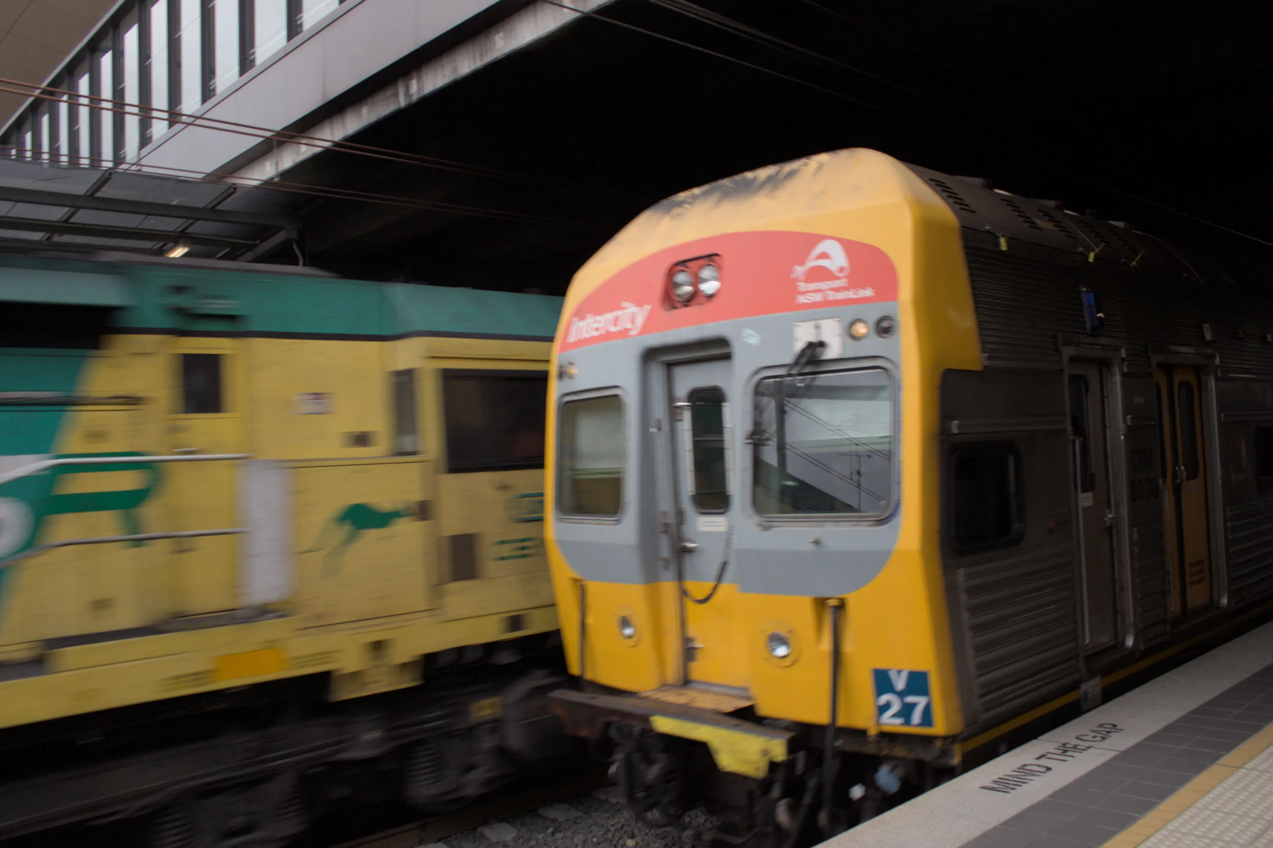 A grey, red and yellow fronted Intercity train pulling into the platform with a fast moving blurry yellow and green "SSR" locomotive moving behind it on the  adjacent track.