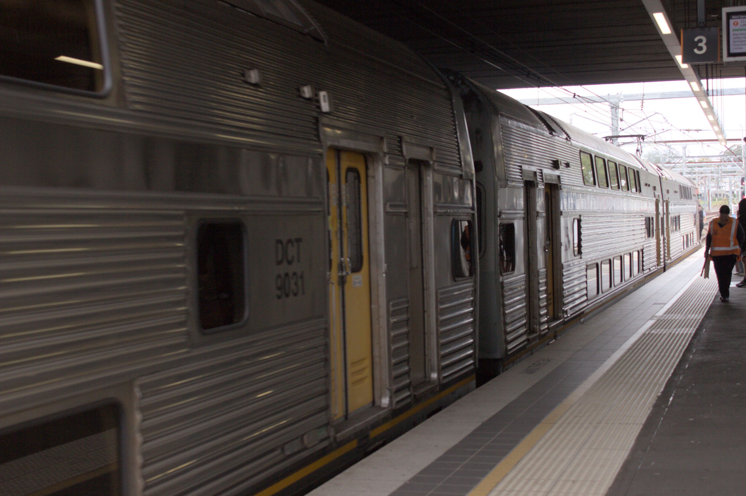 Three silver double-decker train carriages with yellow doors pulling up to a platform, with the closest carriage being identified as "DCT 9031"